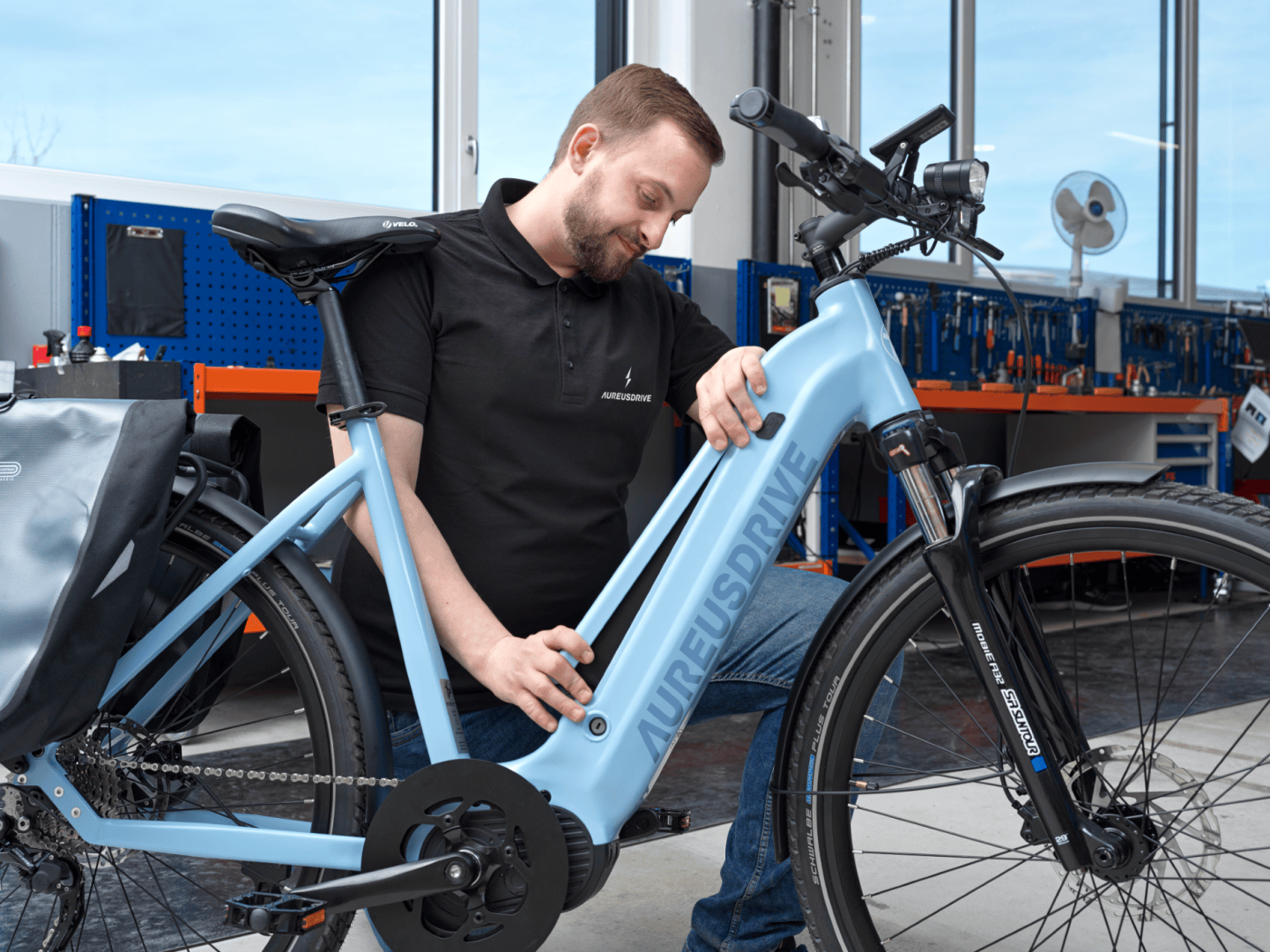 Ein Mann in einem schwarzen Polohemd inspiziert den Rahmen eines hellblauen AUREUSDRIVE-Elektrofahrrads in einer Werkstatt. Im Hintergrund sind Werkbänke und Werkzeuge zu sehen.