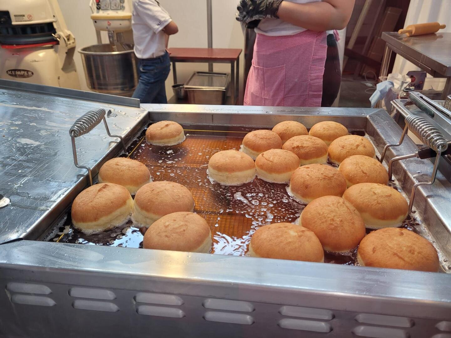 Frisch frittierte runde Krapfen schwimmen in heißem Öl in einer Metallfritteuse in einer Bäckerei. Eine Person in rosa Schürze und Handschuhen steht in der Nähe, im Hintergrund ist die Bäckereiausrüstung zu sehen.