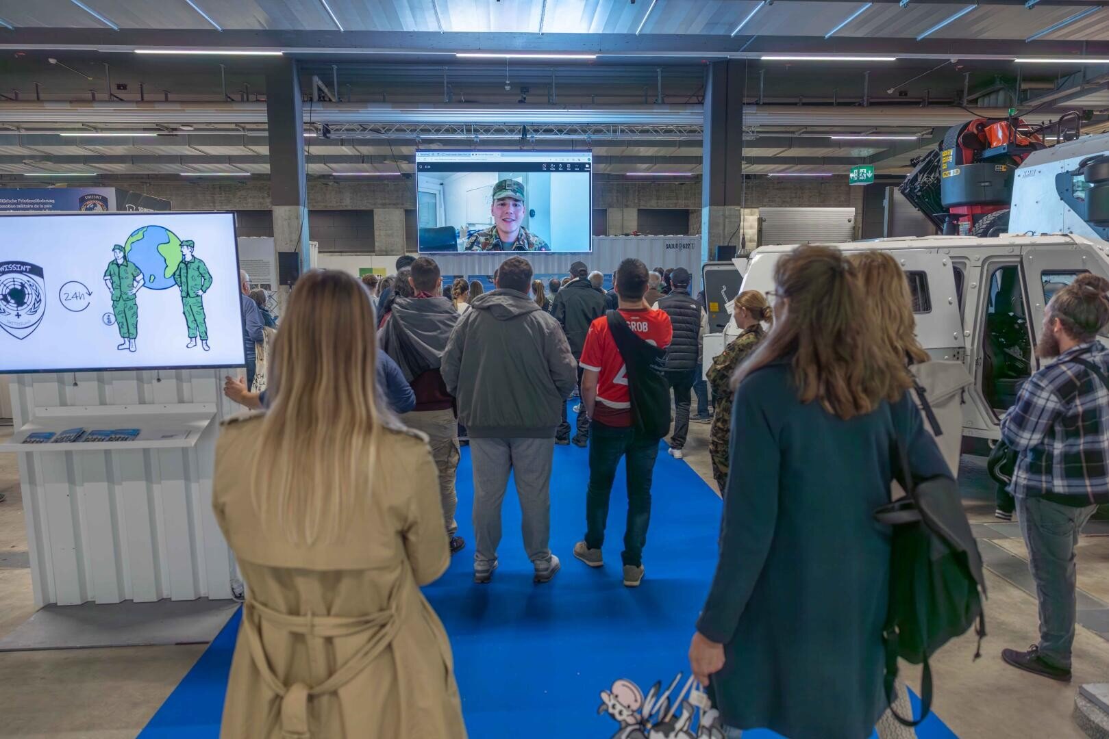 Eine Gruppe von Personen steht auf einem blauen Teppich in einer Halle und sieht einen großen Bildschirm, auf dem eine Person in Militäruniform per Videoanruf spricht. Um das Publikum herum sind Fahrzeuge und Informationsdisplays zu sehen.