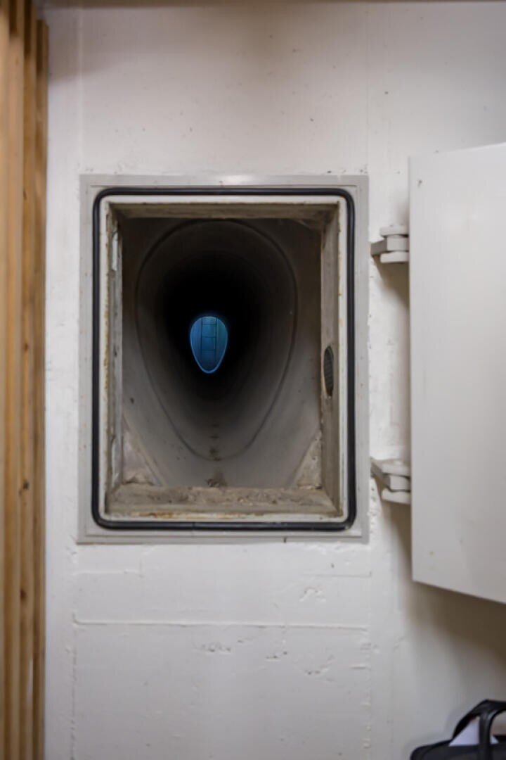 Ein rechteckiger Metalltunnel oder -schacht, der in eine weiße Wand eingebaut ist, mit einer runden Öffnung am anderen Ende, die ein blaues rundes Objekt oder Licht in der Ferne zeigt. Auf der rechten Seite ist eine weiße Schranktür geöffnet.