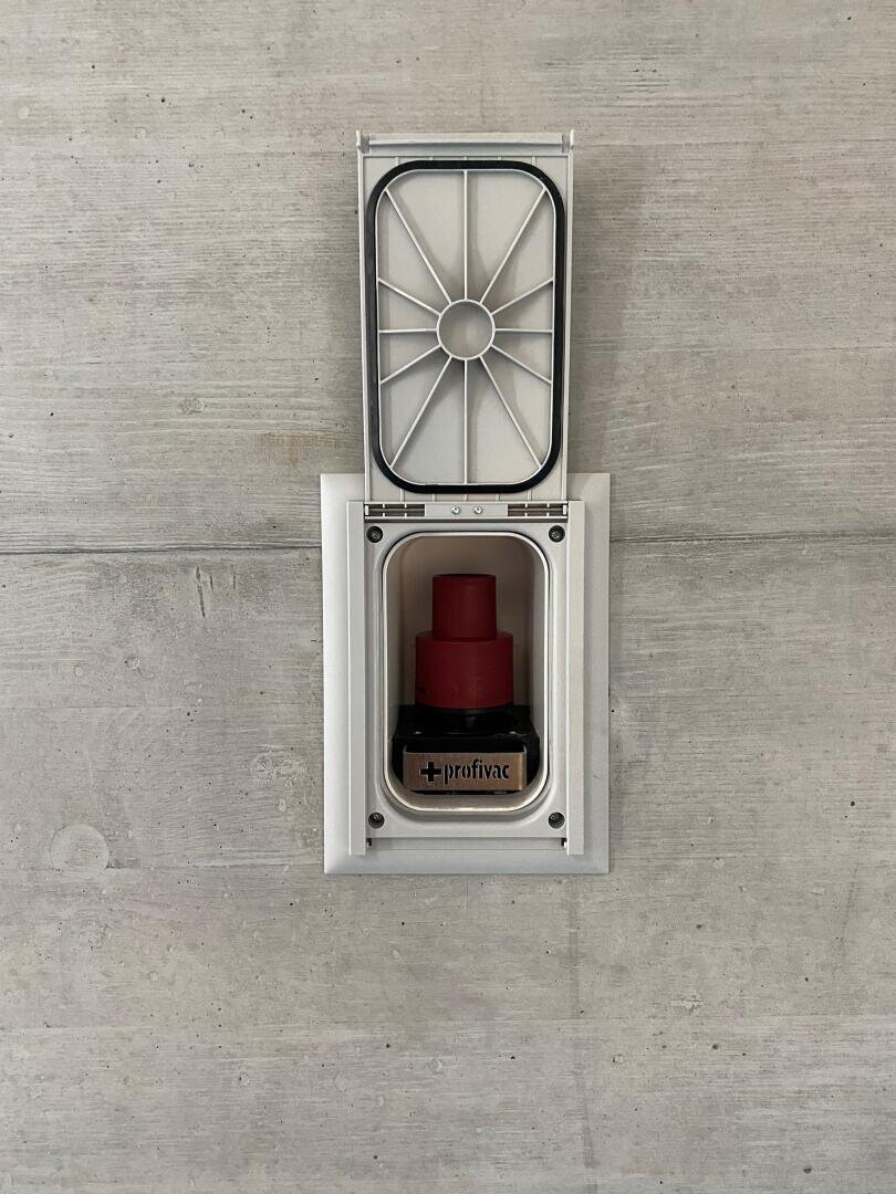 Eine Saugdose mit offenem Deckel zeigt einen rot-schwarzen Profivac-Anschluss vor einem schlichten Betonwandhintergrund.