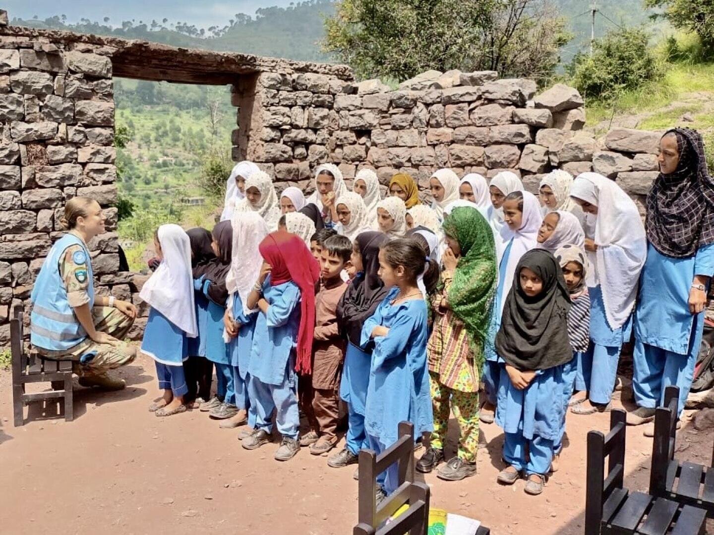 Eine Gruppe von Kindern und Frauen, meist in blauen Uniformen und mit Kopftüchern, steht draußen zwischen den Ruinen eines Steingebäudes und hört einer Person in einer blauen Weste mit einem UN-Abzeichen zu. Im Hintergrund sind grüne Hügel zu sehen.
