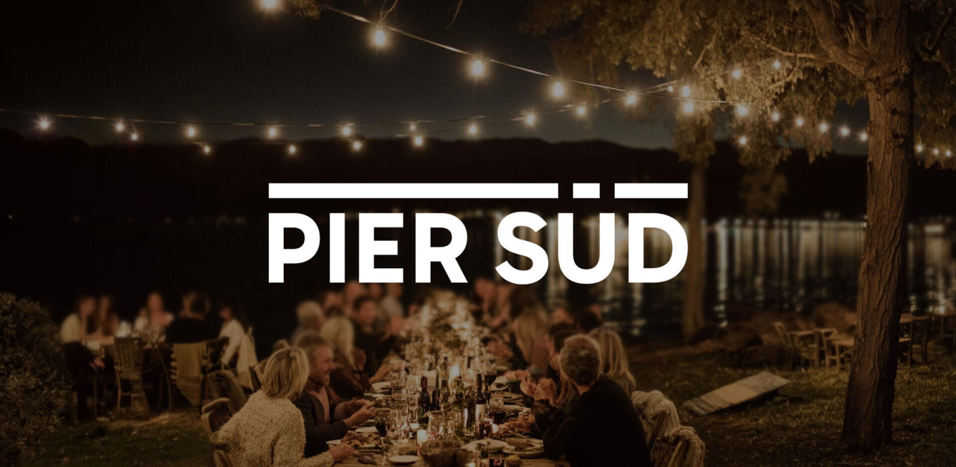 Ein lebhaftes Abendessen im Freien an einem See bei Nacht, mit Lichterketten über dem Kopf und Menschen, die an einem langen Tisch sitzen. Der Schriftzug PIER SÜD erscheint in fetten weißen Buchstaben quer über dem Bild.