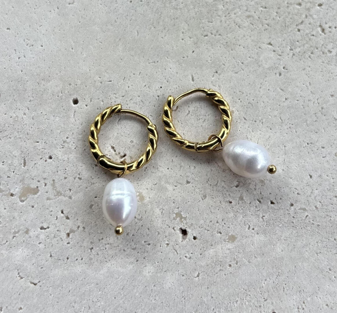 Ein Paar goldene Ohrringe mit gedrehtem Design, an denen jeweils eine einzelne weiße Perle baumelt, auf einer leicht strukturierten Oberfläche.