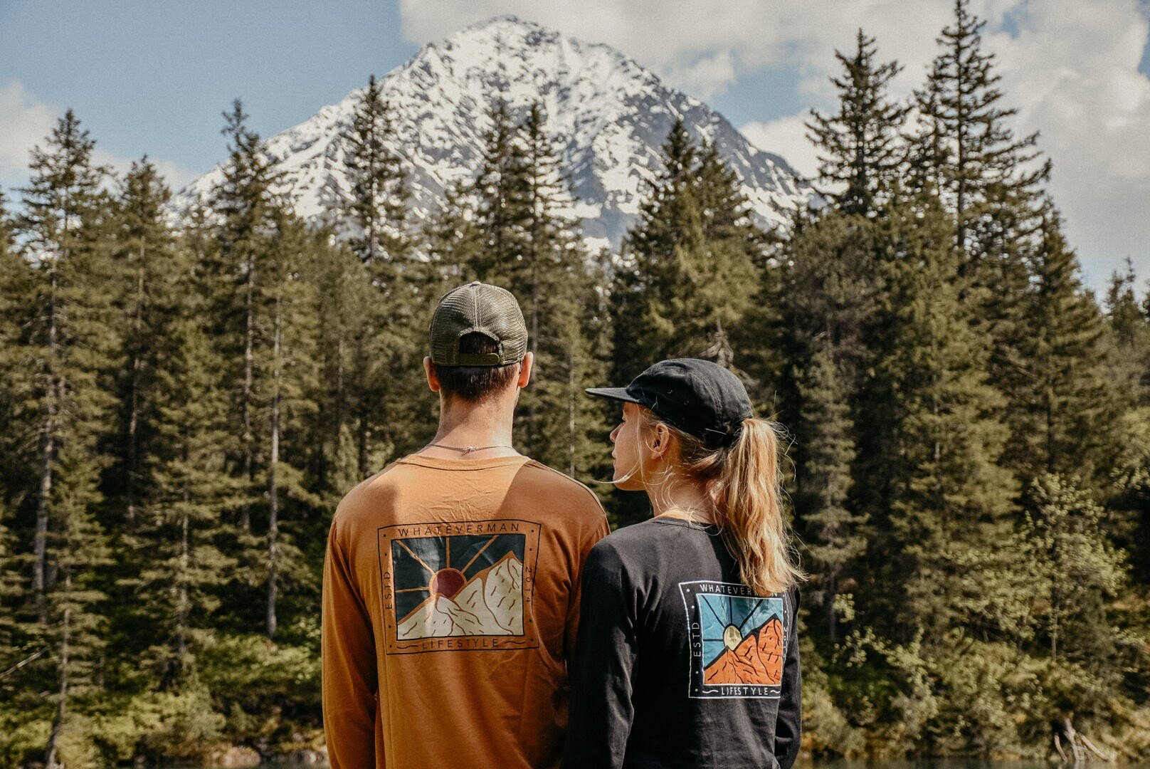 Zwei Personen mit langärmeligen Hemden und Hüten stehen im Freien, mit dem Rücken zur Kamera, und blicken auf einen Kiefernwald und einen schneebedeckten Berg unter einem teilweise bewölkten Himmel.