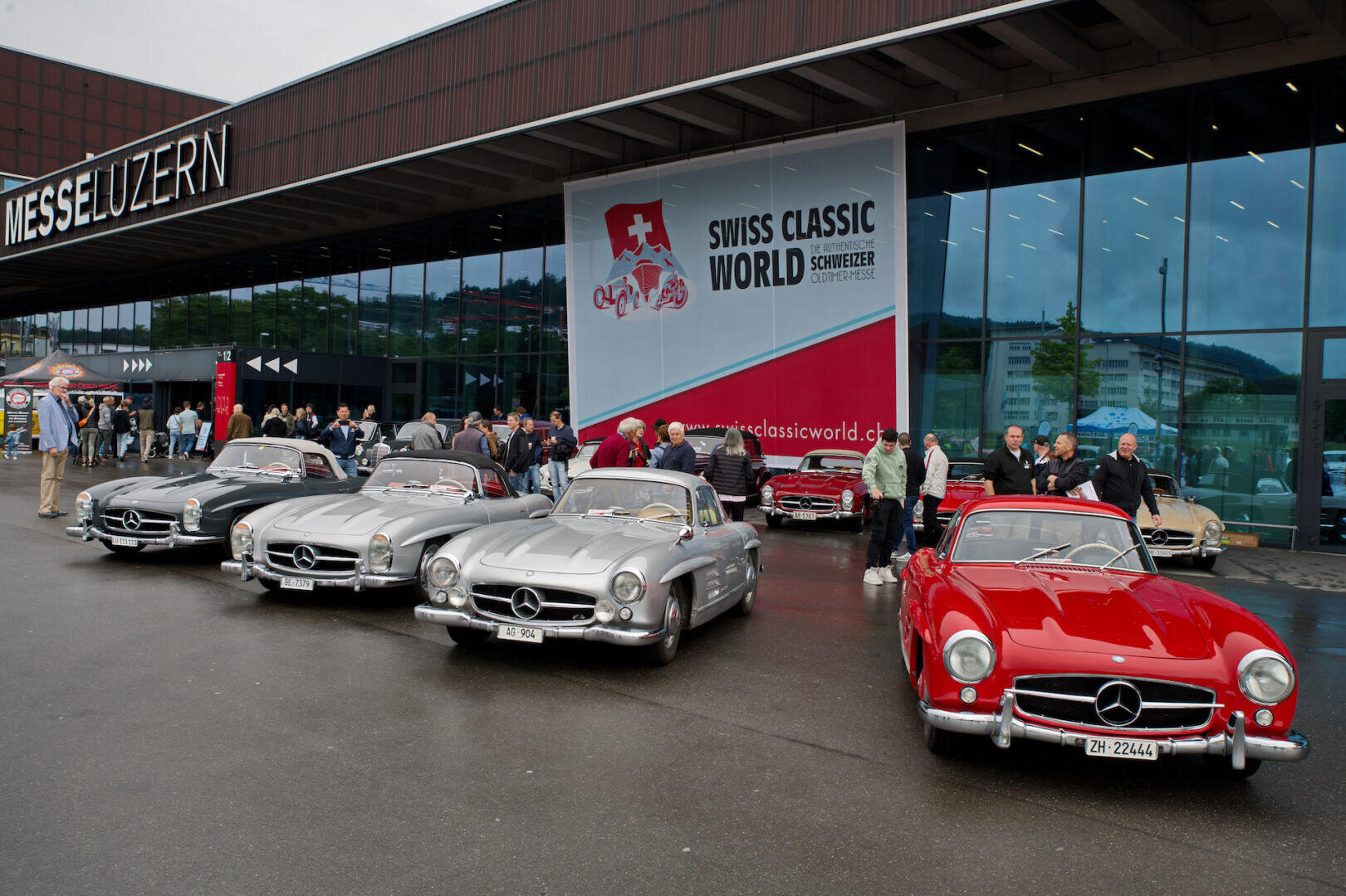 Mehrere Mercedes-Benz-Klassiker, meist silberne und ein roter, sind vor einem modernen Gebäude mit einem Swiss Classic World-Banner geparkt. An einem bewölkten Tag versammelt sich eine Gruppe von Menschen um die Autos.