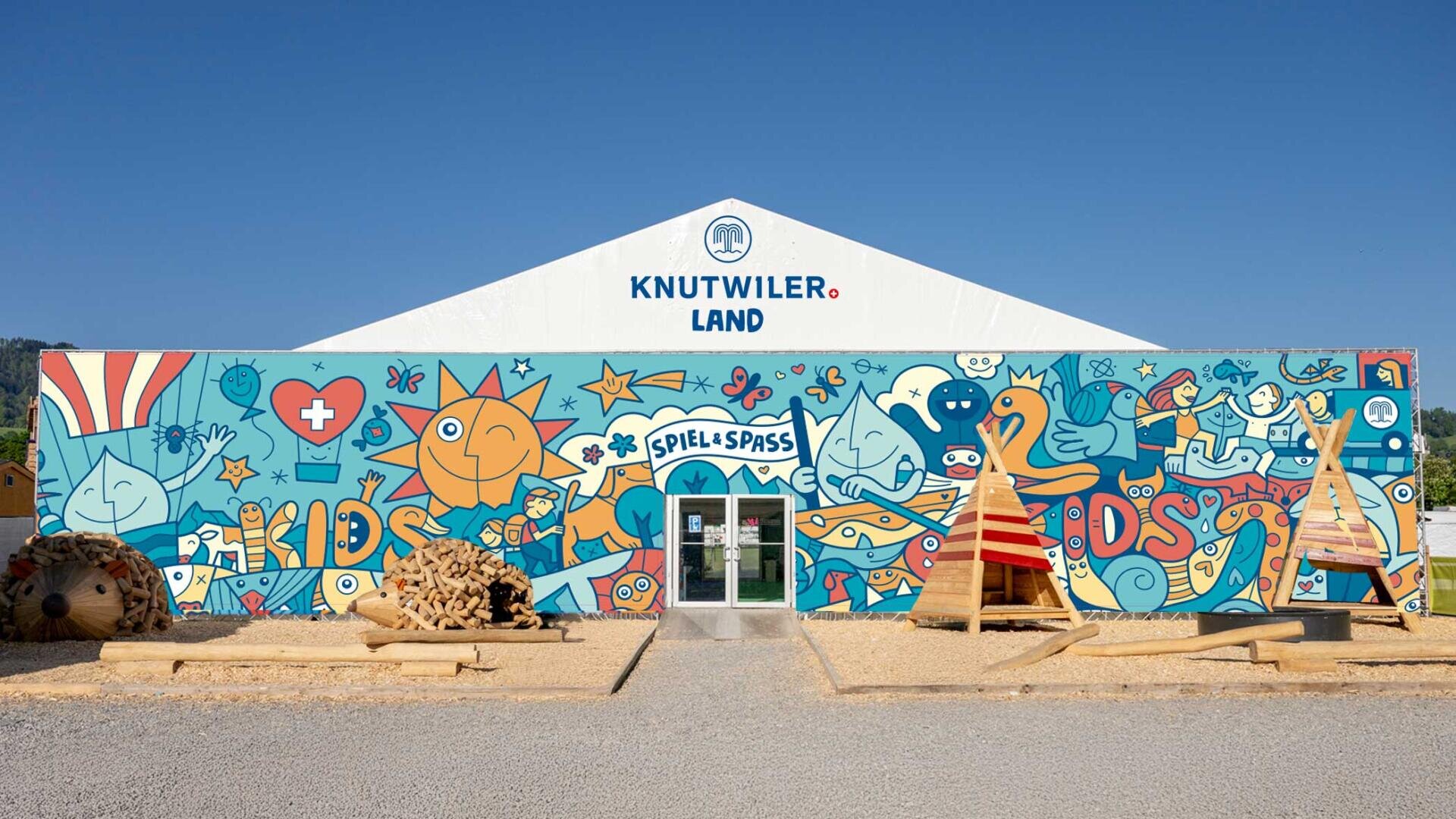Ein farbenfrohes Wandbild auf einem weissen Zelt mit der Aufschrift Knutwiler Land, mit verspielten Illustrationen, fröhlichen Figuren und den Worten Kids und Spiel & Spass, mit Spielgeräten aus Holz im Freien.
