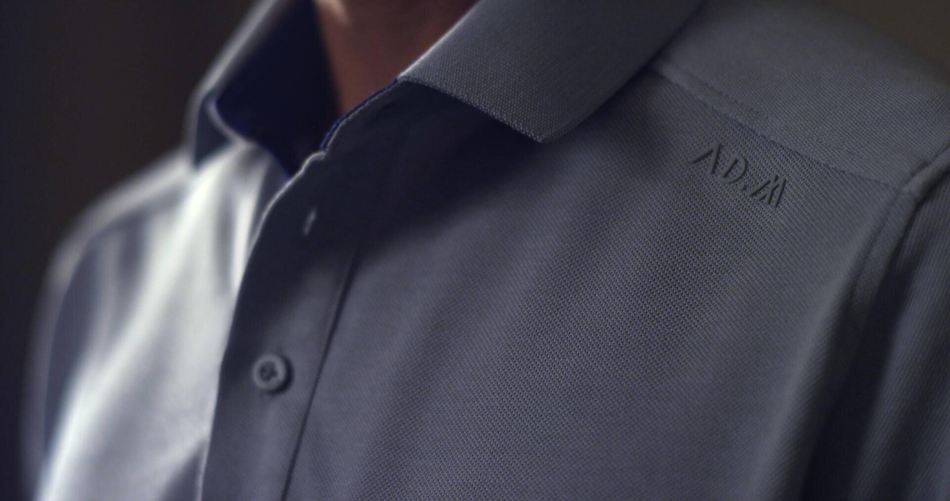 Nahaufnahme einer Person, die ein graues Hemd mit Kragen trägt, bei dem zwei Knöpfe sichtbar sind. Auf dem Hemd ist in der Nähe der Schulter ein dezenter Text mit der Aufschrift ADAY aufgenäht.