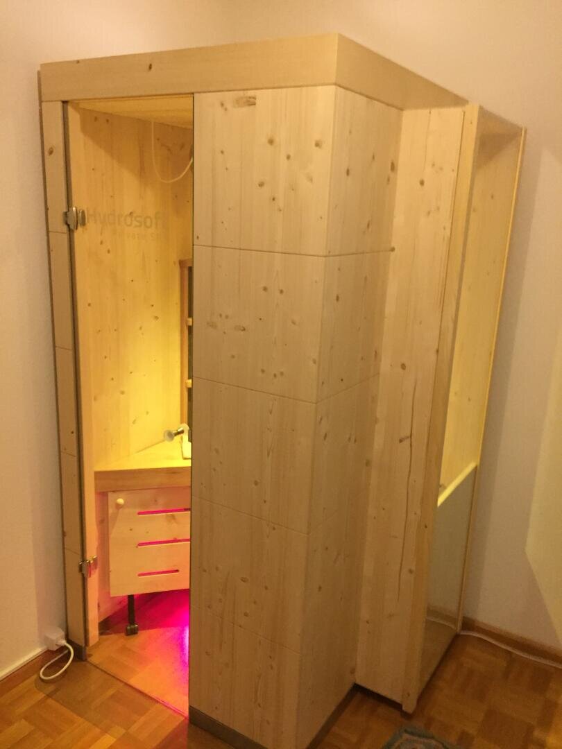 Eine kleine, moderne Holzsaunakabine mit Glastür und Innenbeleuchtung, die in der Ecke eines Raumes mit Parkettboden steht. Der Innenraum ist mit einer Holzbank ausgestattet und leuchtet dezent lila-rosa.