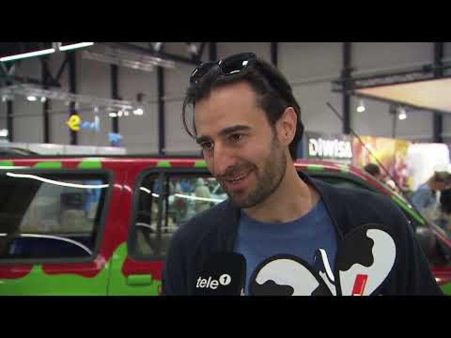 Ein Mann mit Sonnenbrille auf dem Kopf lächelt, während er in einem Innenraum interviewt wird. Er hält ein Mikrofon mit der Aufschrift tele1 in der Hand, und im Hintergrund ist ein buntes Fahrzeug zu sehen.