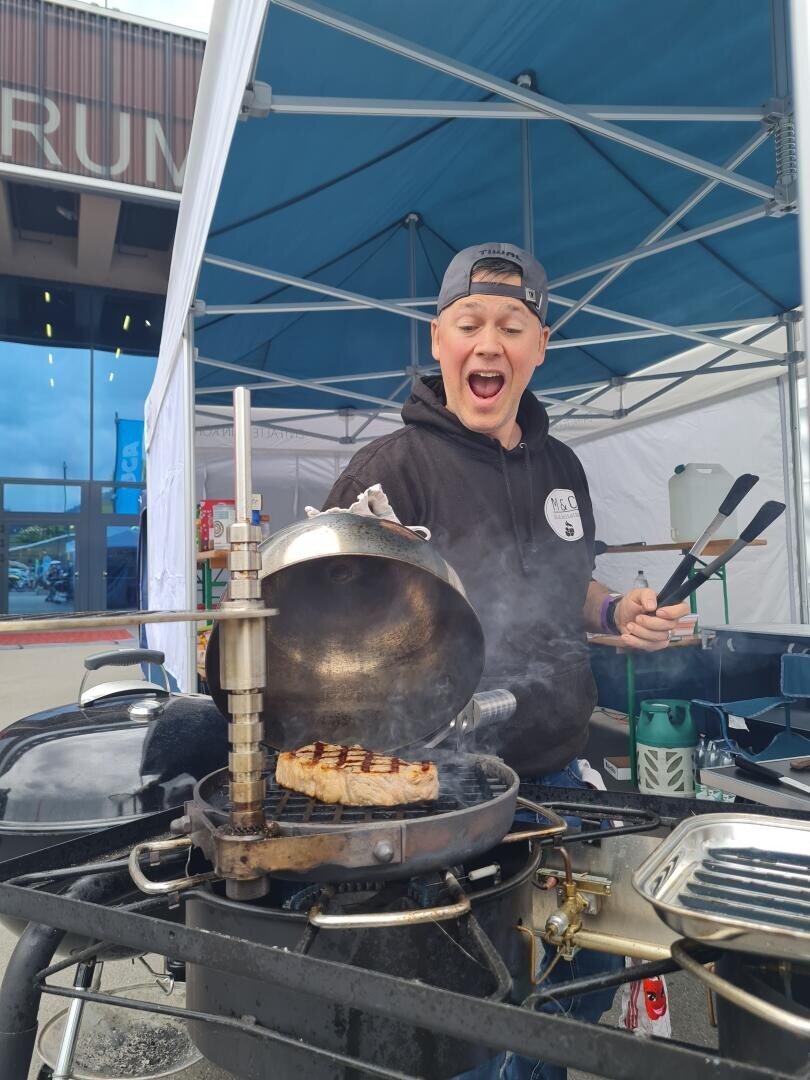 Eine Person mit Kapuzenpulli und Mütze lächelt aufgeregt, während sie unter einem Zeltdach Fleisch grillt und eine Zange hält, aus der Rauch aufsteigt.