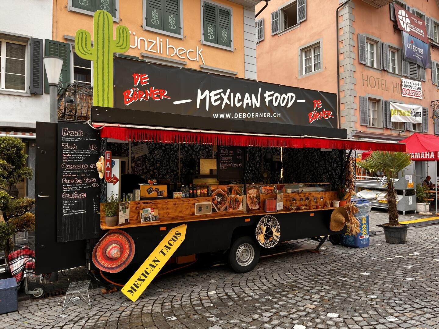 Ein bunter mexikanischer Imbisswagen mit einem großen Schild mit der Aufschrift "Mexican Food" ist auf einer Kopfsteinpflasterstraße geparkt. Auf dem Wagen sind Speisekarten, Dekorationen, Sombreros und eine große Kaktusfigur zu sehen, während im Hintergrund Gebäude zu sehen sind.