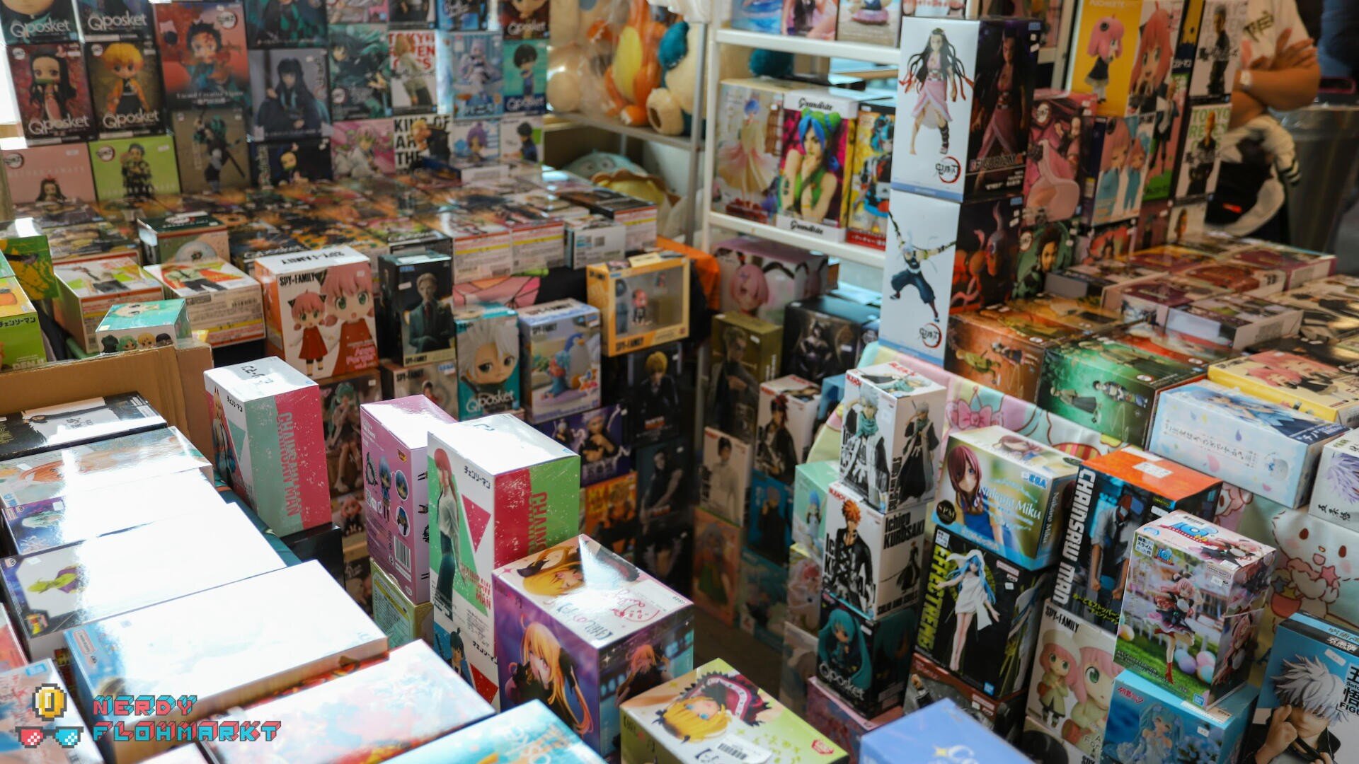 Eine überfüllte Auslage mit bunten Anime-Figuren und -Modellboxen, die auf Tischen und in Regalen auf einer Messe oder in einem Geschäft gestapelt sind und verschiedene Figuren und Serien in farbenfrohen Verpackungen zeigen.