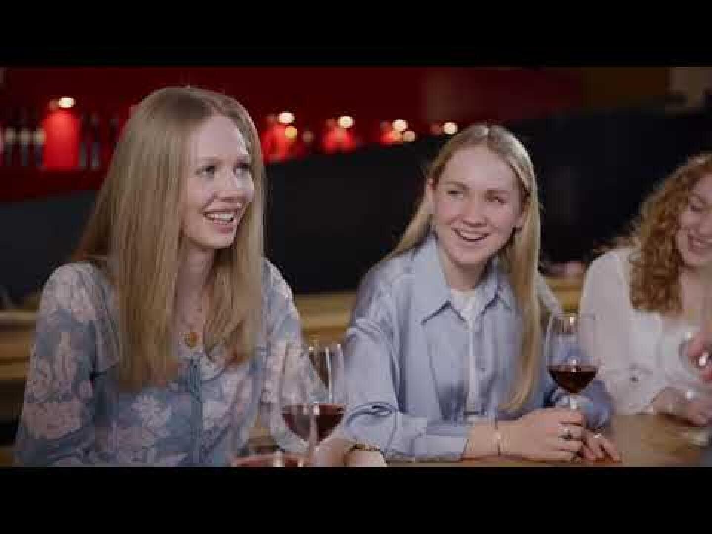 Drei junge Frauen sitzen an einem Tisch, lächeln und unterhalten sich, während sie Gläser mit Rotwein halten. Der Hintergrund ist warm beleuchtet und suggeriert eine entspannte, gesellige Atmosphäre.