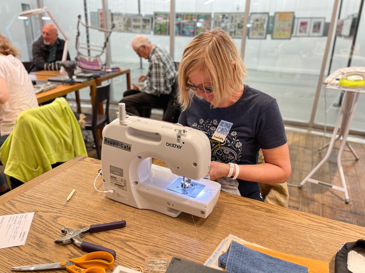 Eine Frau arbeitet mit einer Brother-Nähmaschine an einem Tisch in einem hellen, gemeinschaftlichen Arbeitsbereich. Andere Personen arbeiten an den benachbarten Tischen. Werkzeuge und Stoffe liegen auf dem Tisch, und im Hintergrund sind Kunstwerke ausgestellt.