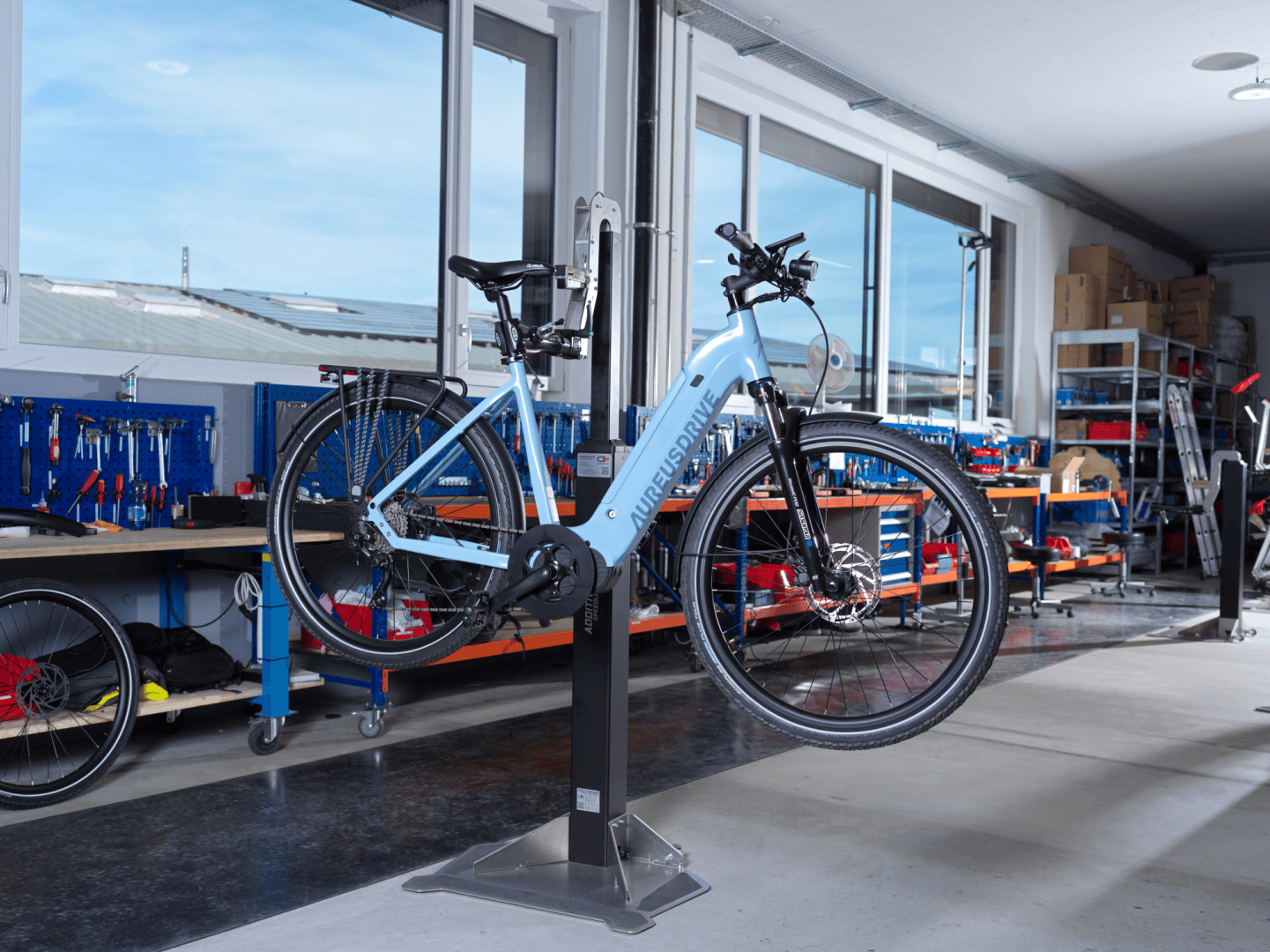 Ein hellblaues Elektrofahrrad steht auf einem Reparaturständer in einer Werkstatt, umgeben von Werkzeugen, Werkbänken und Ersatzteilen, mit großen Fenstern, die Tageslicht hereinlassen.