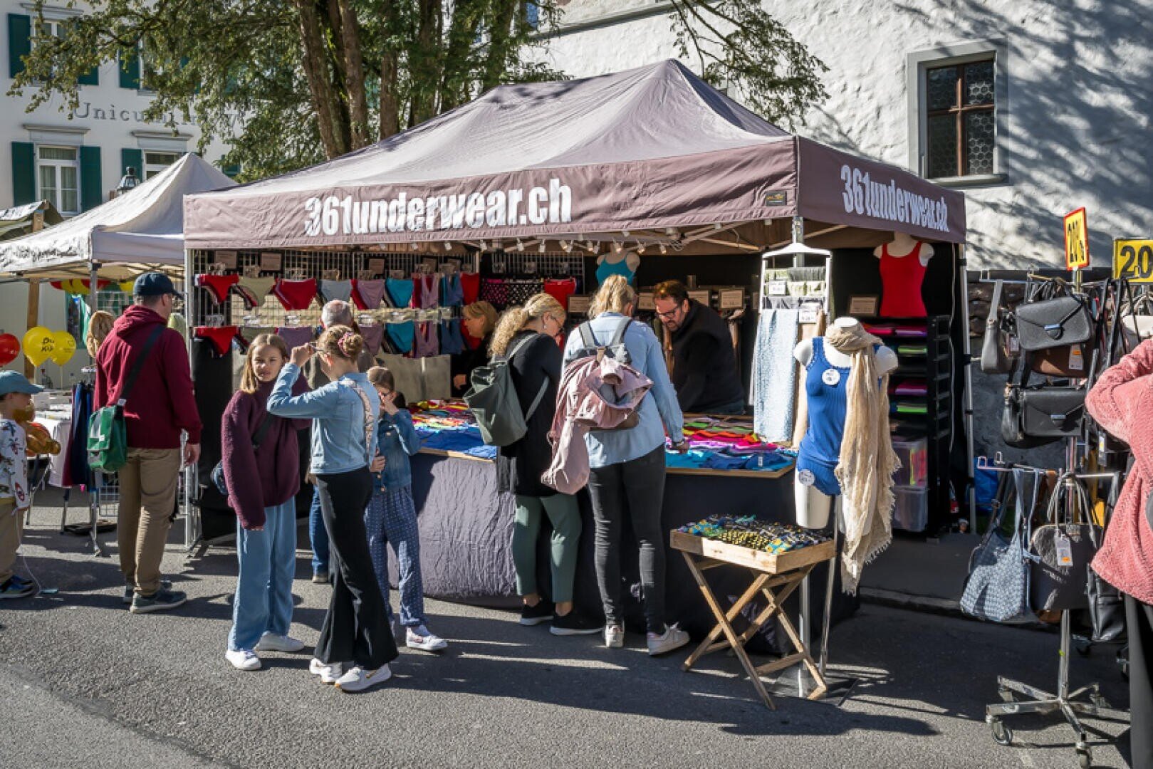 Eine Gruppe von Menschen stöbert an einem sonnigen Tag an einem Marktstand unter einem Zelt mit der Aufschrift 36underwear.ch in Kleidung und Accessoires. Verschiedene Kleidungsstücke und Taschen sind auf Tischen und Regalen angeordnet.