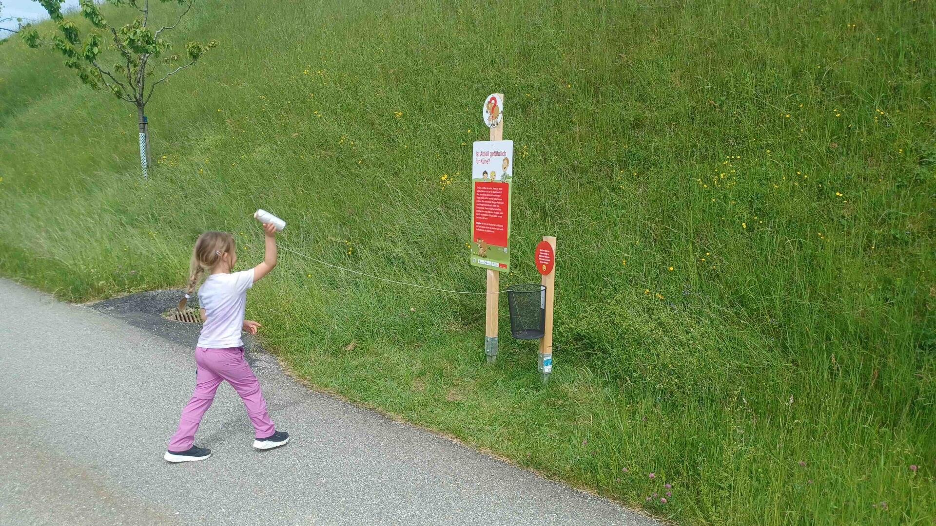 Ein junges Mädchen in rosafarbener Hose und weißem Hemd hält sich einen an einer Schnur befestigten Becher ans Ohr und beschäftigt sich auf einem mit Wildblumen und einem kleinen Baum gesäumten Grasweg mit einer Outdoor-Aktivitätstafel.