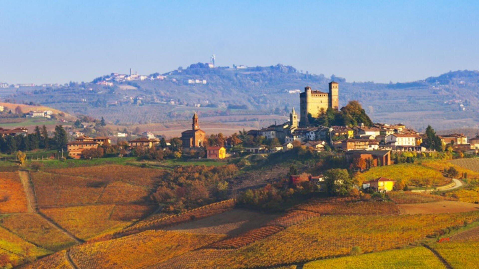 Ein malerisches Dorf auf einem Hügel mit einem Schloss, einer Kirche und Häusern, umgeben von bunten Weinbergen und sanften Hügeln unter einem klaren blauen Himmel.