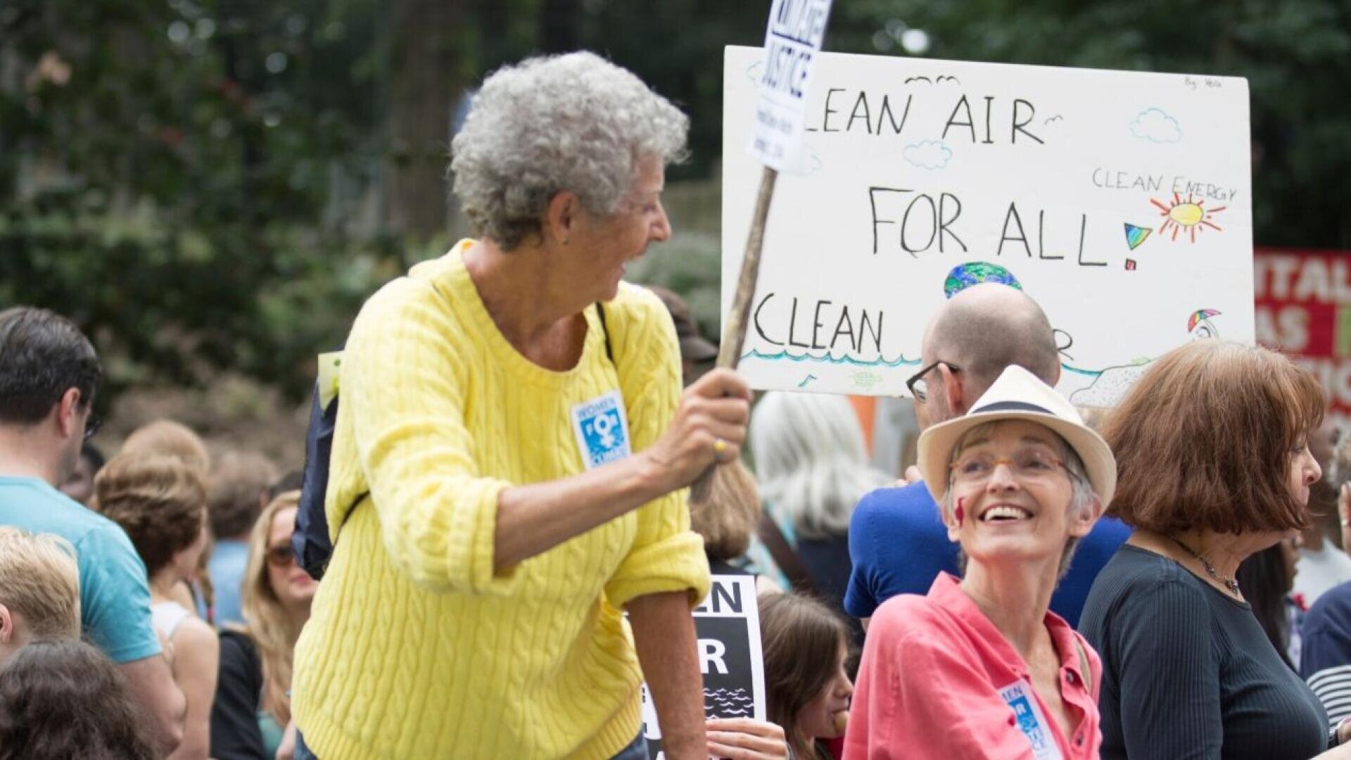 Eine Gruppe von Menschen bei einer Kundgebung im Freien, mit zwei älteren Frauen im Vordergrund, die Schilder halten. Auf einem Schild steht "SAUBERE LUFT FÜR ALLE". Die Stimmung scheint positiv und lebhaft zu sein.