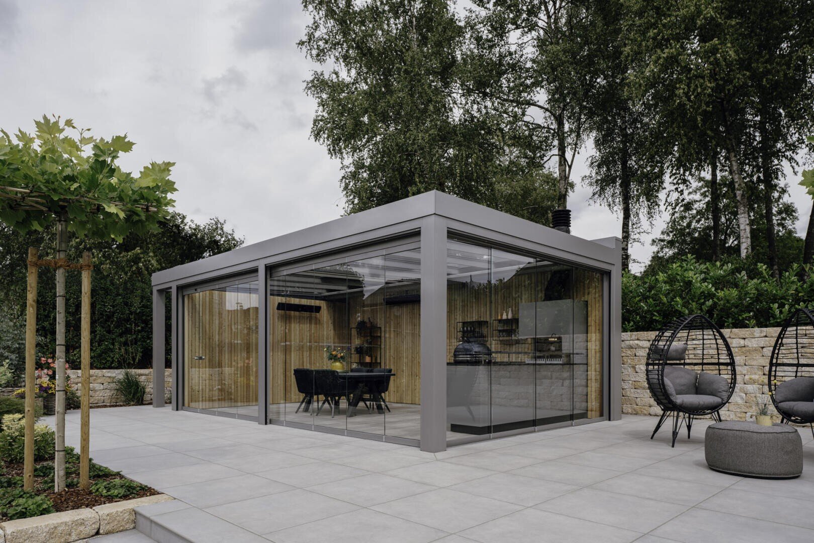 Ein modernes, verglastes Gartenzimmer mit einem Esstisch und Stühlen im Inneren, das auf einer gefliesten Terrasse steht. Das umgebende Grün, die Bäume und die Steinmauern schaffen eine ruhige Umgebung im Freien, in der sich auch Hängesessel befinden.