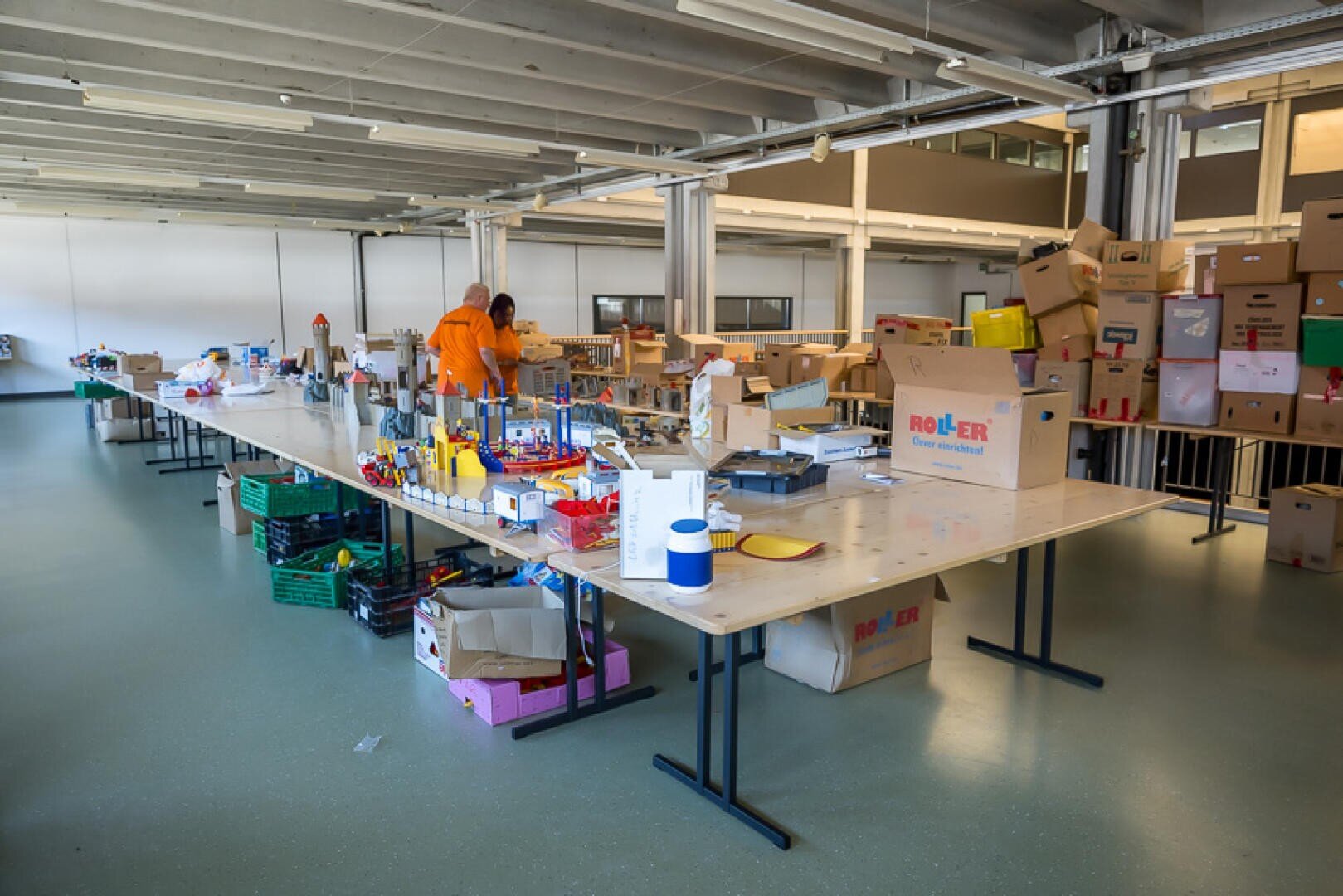 Ein geräumiger Raum mit langen Tischen, die mit verschiedenen Materialien und Kartons bedeckt sind. Zwei Personen in orangefarbenen Hemden arbeiten inmitten vieler gestapelter Kartons und verstreuter Gegenstände, was auf eine organisierte Sortier- oder Verpackungstätigkeit schließen lässt.