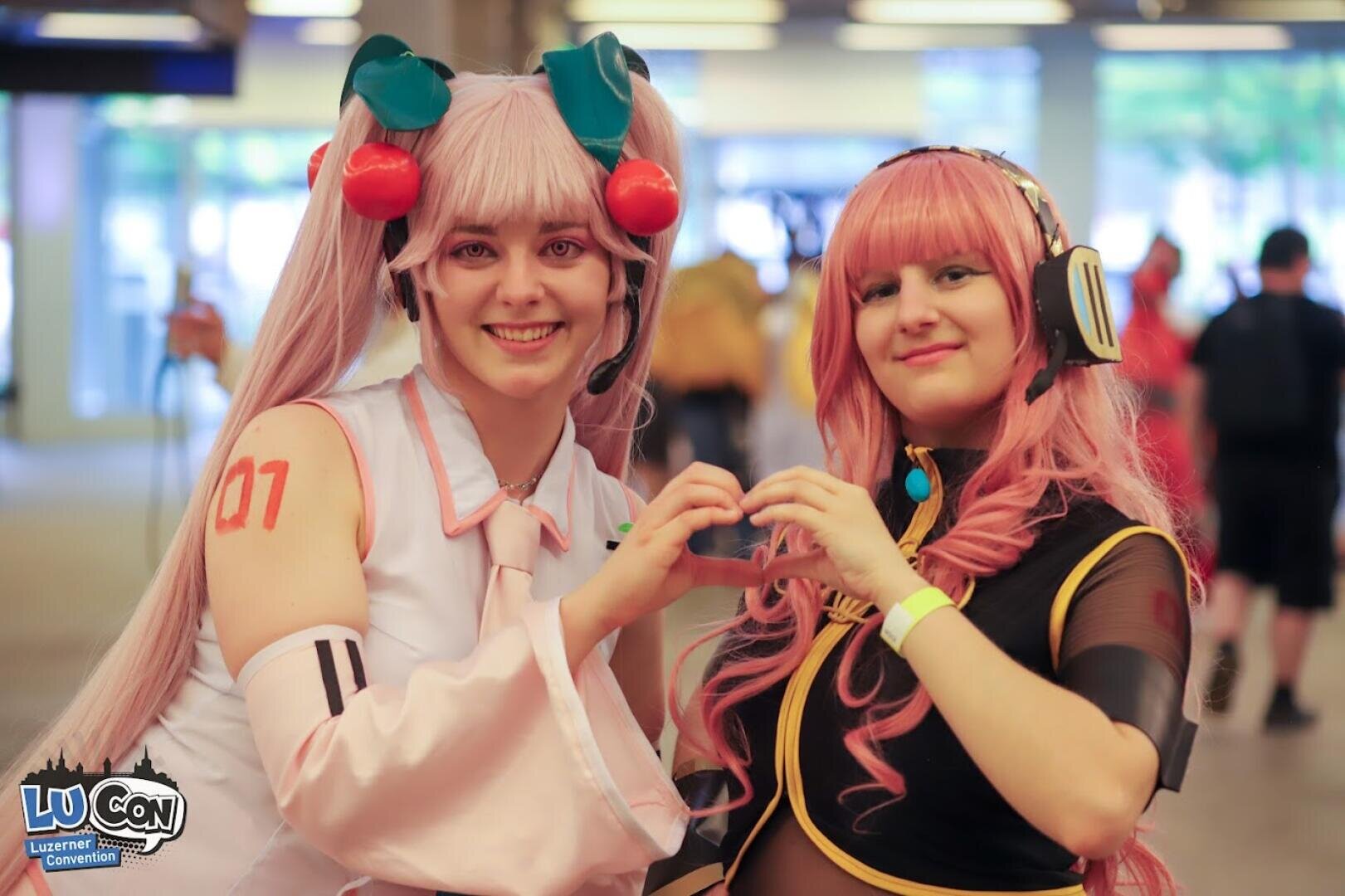 Zwei Personen in farbenfrohem Anime-Cosplay posieren zusammen auf einer Convention und bilden mit ihren Händen eine Herzform. Die eine trägt lange rosa Zwillingsschwänze und Haarschmuck, die andere hat lange rosa Haare, Kopfhörer und ein schwarzes Outfit.