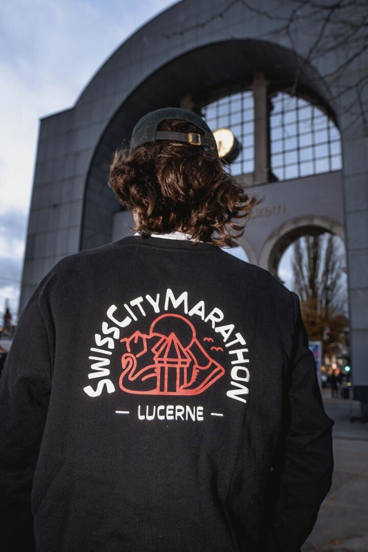 Eine Person mit langen Haaren und einer nach hinten gerichteten Mütze steht vor einem gewölbten Gebäude und trägt ein schwarzes Sweatshirt mit der Aufschrift Swiss City Marathon Lucerne und einer roten Grafik auf dem Rücken.