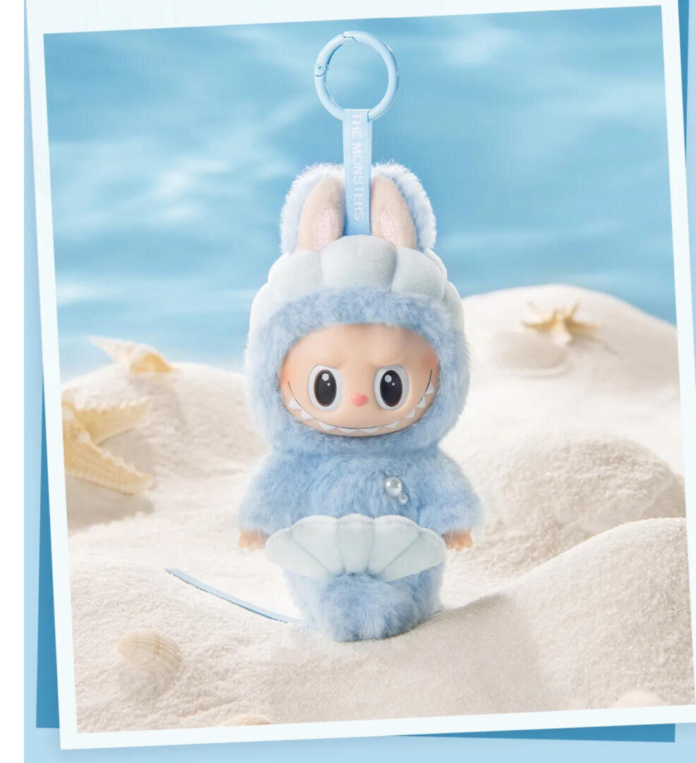 Eine niedliche Puppe mit großen Augen, bekleidet mit einem flauschigen blauen Häschenkostüm und einer Muschel in der Hand, steht auf weißem Sand mit Seesternen im Hintergrund. Am Kopf der Puppe ist ein Schlüsselbund befestigt. Die Szene hat eine weiche, skurrile Ausstrahlung.