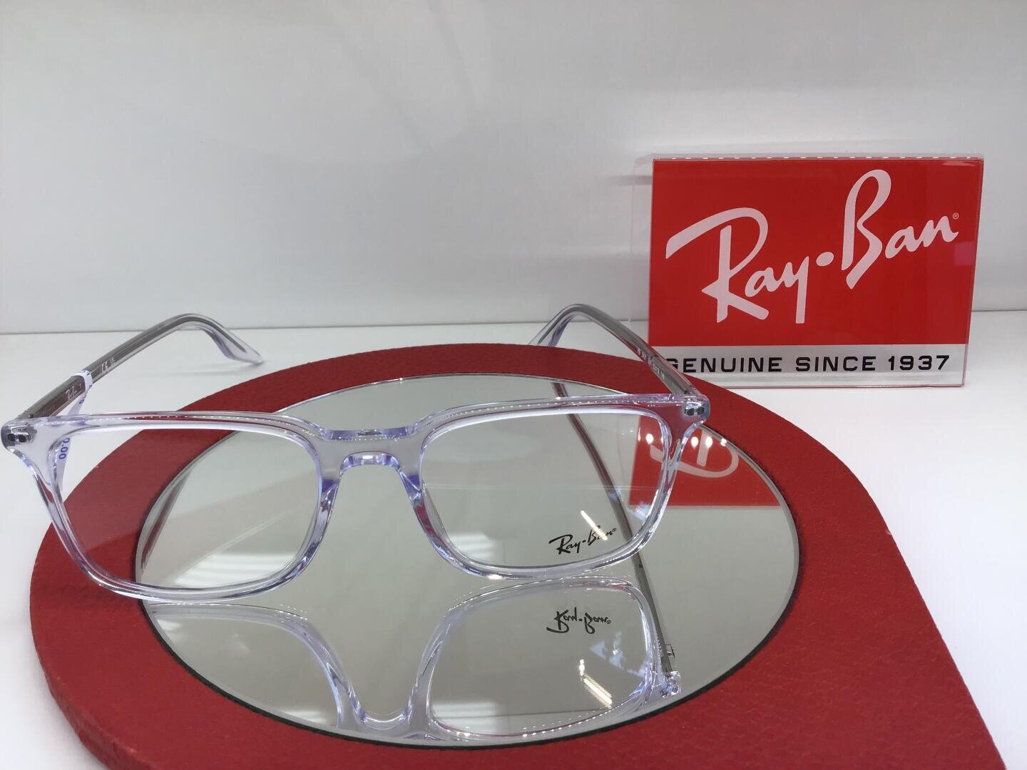 Eine klare Ray-Ban-Brille ruht auf einem runden Spiegel mit rotem Rand. Im Hintergrund ist ein rot-weißes Ray-Ban-Zeichen mit der Aufschrift "Genuine since 1937" zu sehen.