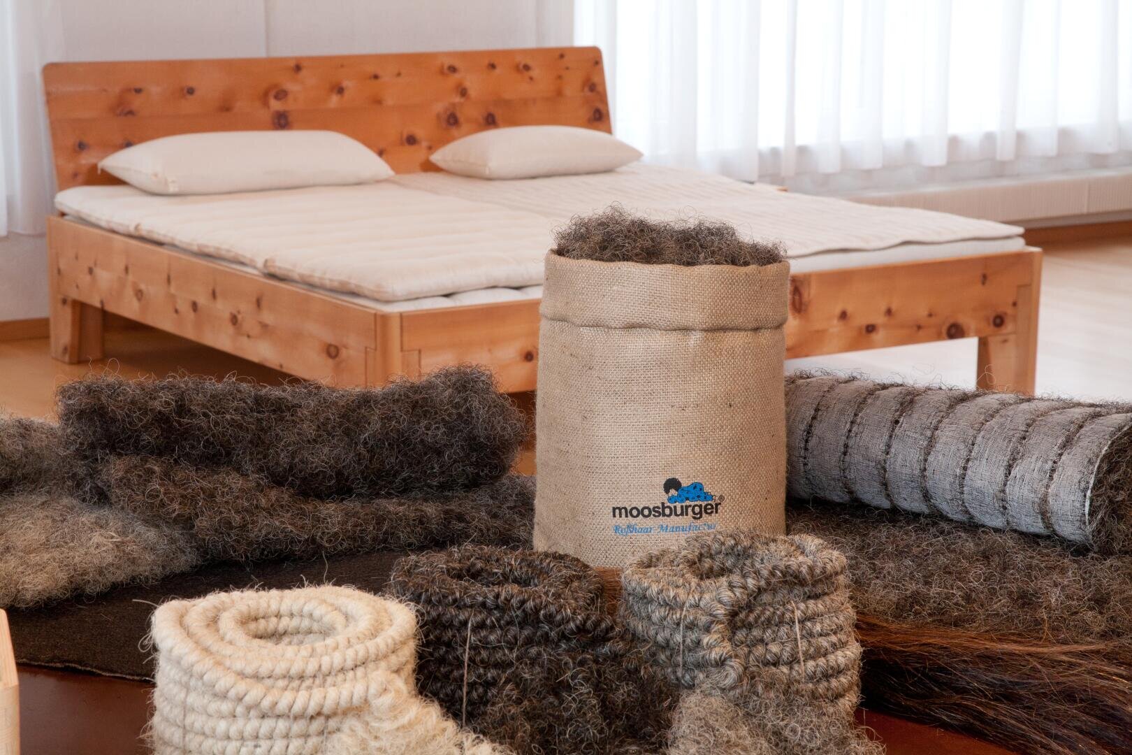 Ein Holzbett mit zwei Kissen und einer Matratze steht in einem hellen Raum. Im Vordergrund befinden sich Matratzenmaterialien aus Naturfasern, Rollen und ein Sack aus Sackleinen mit der Aufschrift Moosburger. Die Szene deutet auf umweltfreundliche Bettwaren hin.