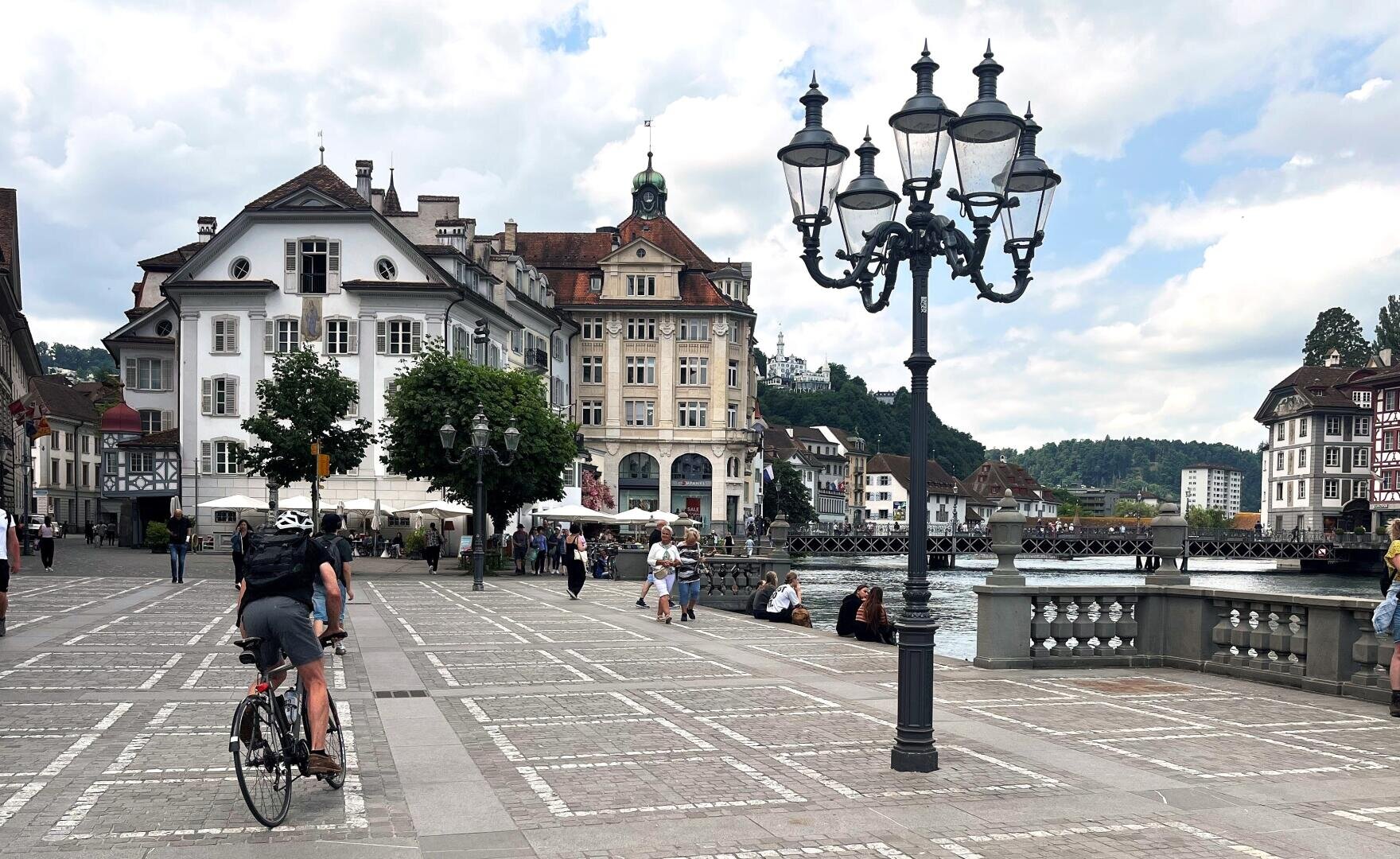 Ein Radfahrer fährt über einen Platz mit Kopfsteinpflaster, der von historischen Gebäuden, Straßenlaternen und Bäumen in der Nähe eines Flusses gesäumt ist, wo Menschen spazieren gehen und auf Bänken sitzen, während der Himmel bewölkt ist.
