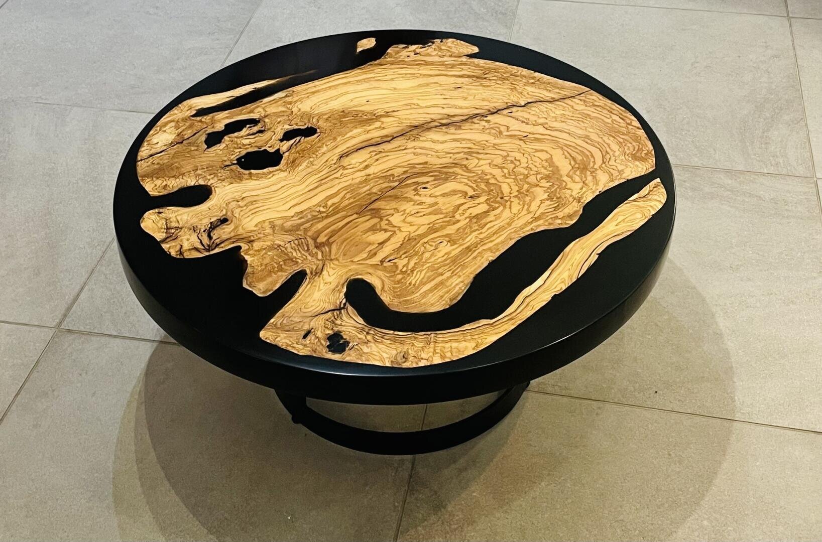 Ein runder Couchtisch mit schwarzem Sockel und schwarzer Platte, der durch eine auffällige Holzintarsie mit natürlichen, wirbelnden Maserungen und unregelmäßigen Kanten ein künstlerisches und organisches Aussehen erhält. Der Tisch steht auf einem hellen Fliesenbelag.