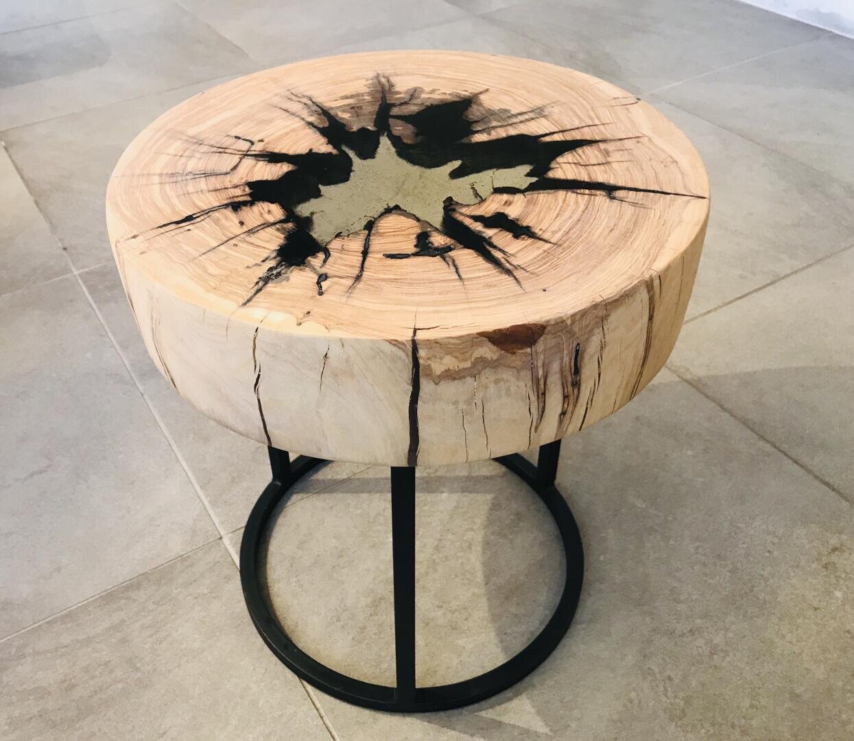 Ein runder Holzhocker mit einem natürlichen Rissmuster, der oben mit schwarzem Harz gefüllt ist, getragen von schwarzen Metallbeinen mit einem runden Sockel, der auf einem gefliesten Boden steht.