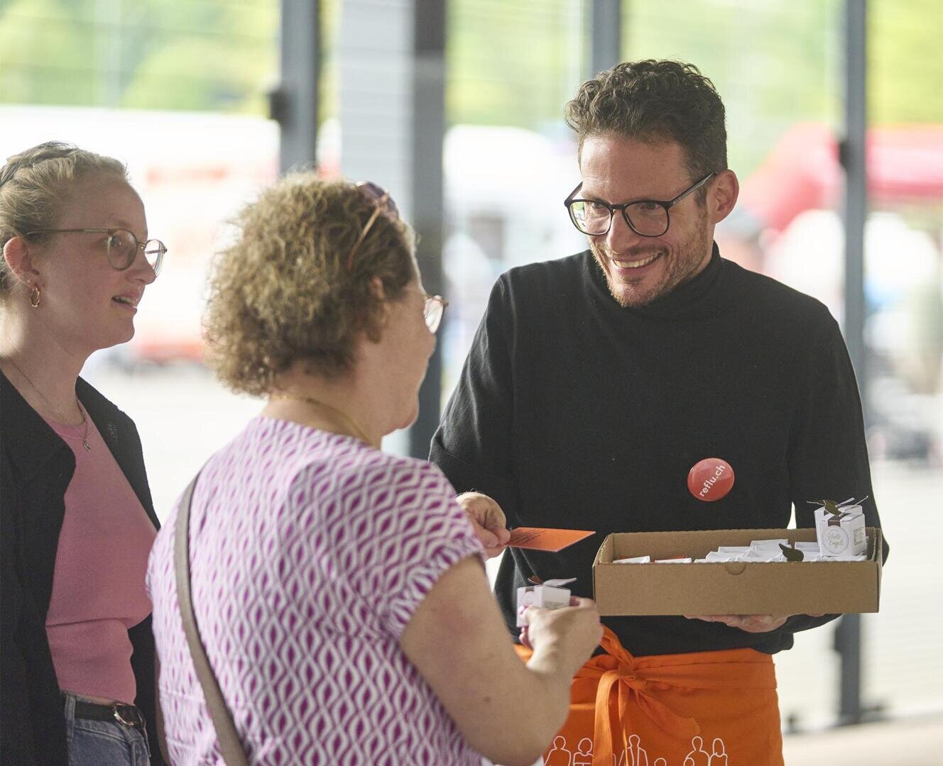 Ein lächelnder Mann mit Brille und schwarzem Hemd reicht zwei Frauen in einem Haus kleine Gegenstände aus einer Schachtel; die eine Frau greift nach einem Gegenstand, während die andere zusieht.