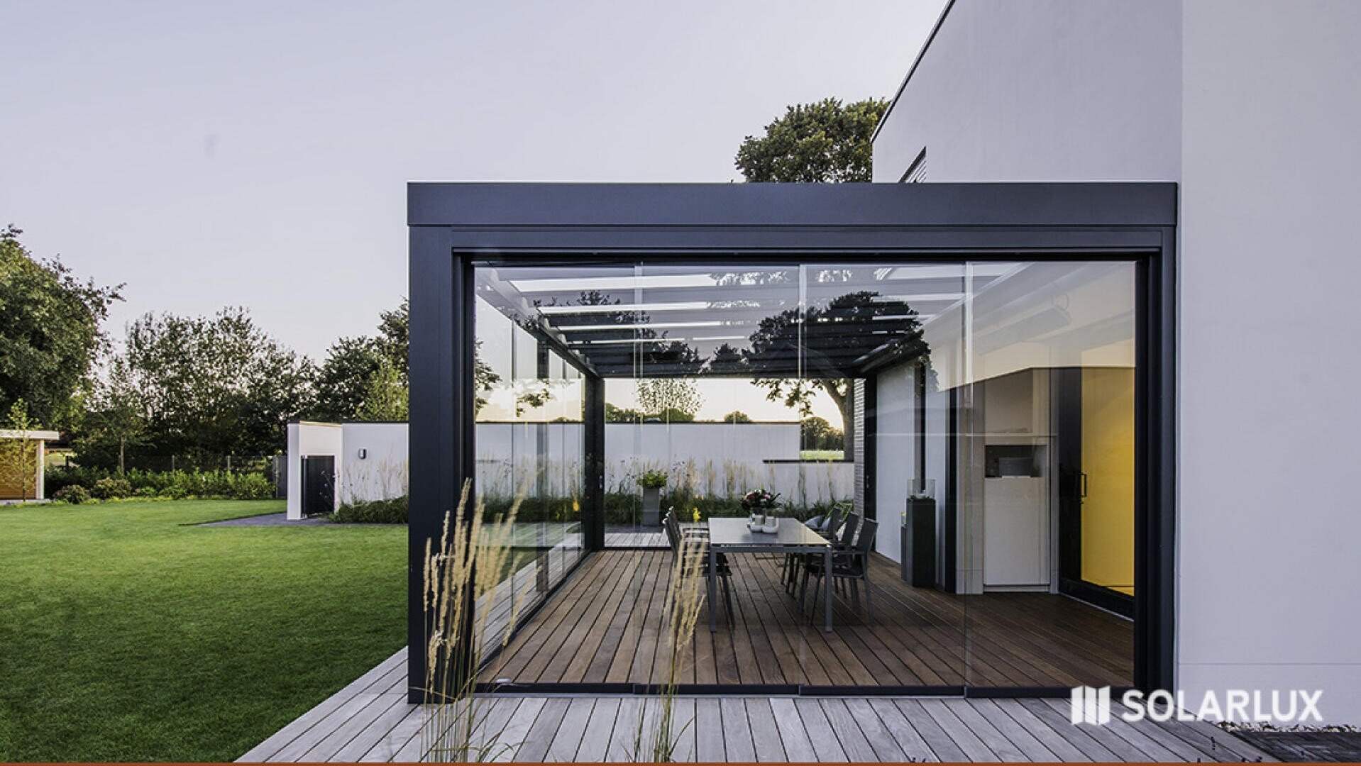 Ein moderner Raum mit Glaswänden und einem Esstisch öffnet sich zu einem üppigen grünen Garten. Die Struktur weist schlichte schwarze Rahmen und einen Holzboden auf. Das Solarlux-Logo ist in der unteren rechten Ecke zu sehen.