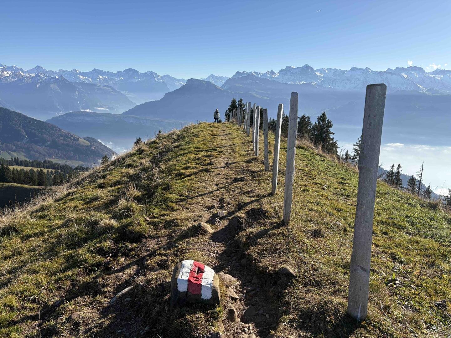 Ein felsiger, mit Holzpfählen gesäumter Pfad führt entlang eines Bergkamms; in der Ferne steht ein Wanderer. Im Hintergrund sind schneebedeckte Gipfel und ein strahlend blauer Himmel zu sehen. Im Vordergrund befindet sich ein Stein mit einer rot-weißen Wegmarkierung.