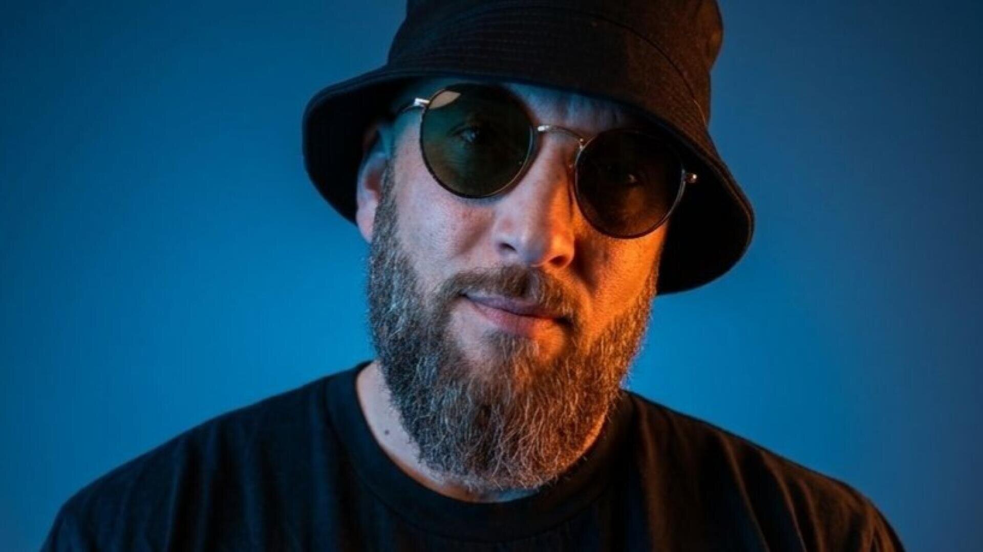 Ein Mann mit Vollbart und Schnurrbart trägt einen schwarzen Eimerhut, eine runde Sonnenbrille und ein schwarzes Hemd vor einem blauen Hintergrund.