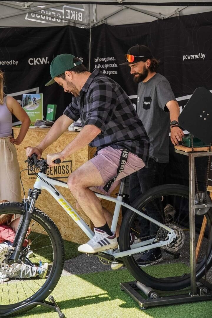 Ein Mann mit Mütze und kurzer Hose tritt an einem Stand im Freien in die Pedale eines stationären Fahrrads, während ein anderer Mann mit Sonnenbrille lächelnd daneben steht. Eine Frau steht teilweise sichtbar an der Theke des Standes.