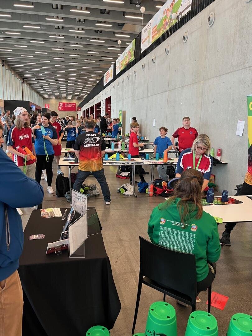 Menschen verschiedenen Alters nehmen an einem Becherstapelwettbewerb in einer großen Halle teil. Einige stapeln Becher an Tischen, während andere zusehen oder warten, bis sie an der Reihe sind. In der geschäftigen Szene sind Schilder, Banner und Geräte zu sehen.