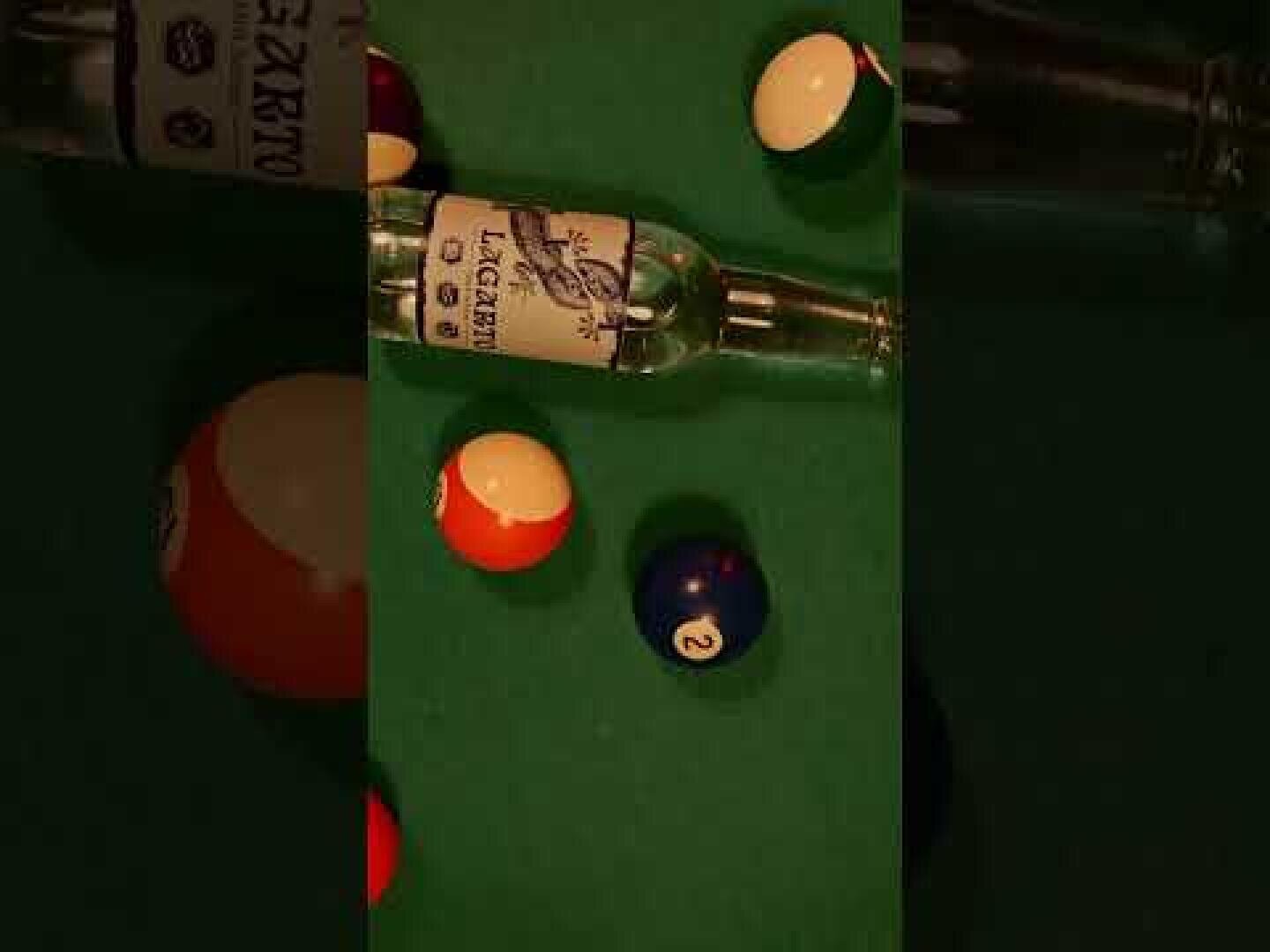 Ein Billardtisch mit bunten Billardkugeln und einer leeren Glasflasche, die in der Mitte auf der Seite liegt. Die Ansicht ist leicht schräg, und oben rechts ist ein Teil einer anderen Flasche zu sehen.