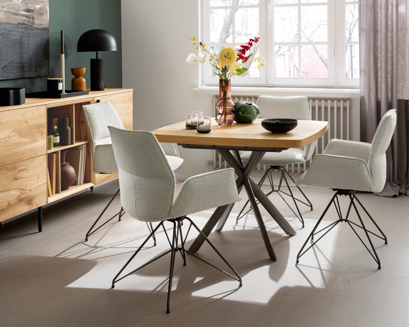 Ein modernes Esszimmer mit einem Holztisch, vier weiß gepolsterten Stühlen und einer Holzanrichte. Auf dem Tisch und der Anrichte befinden sich Dekorationsartikel und Blumen. Ein großes Fenster lässt natürliches Licht herein.