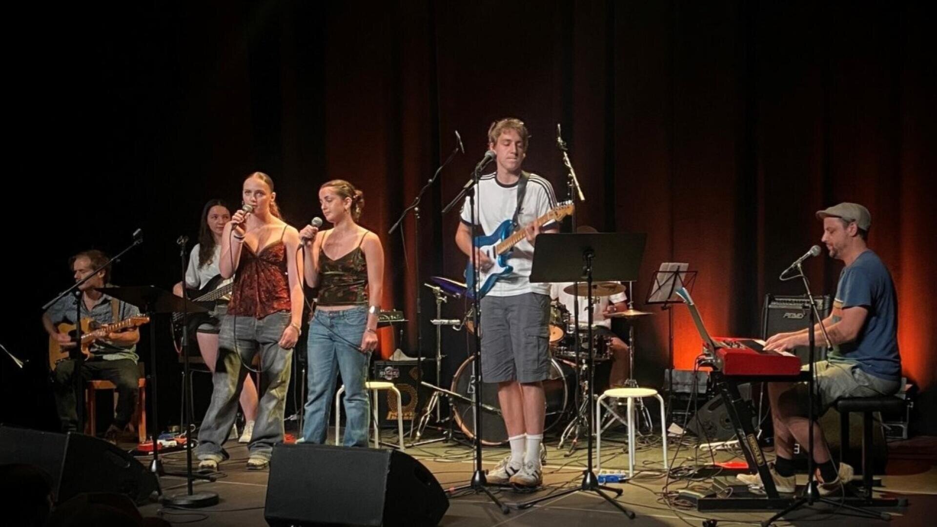 Eine sechsköpfige Band tritt auf der Bühne mit Gitarren, Keyboard, Schlagzeug und Mikrofonen auf. Zwei Frauen und ein Mann singen, während die anderen Instrumente spielen. Warmes Bühnenlicht erhellt die Gruppe.