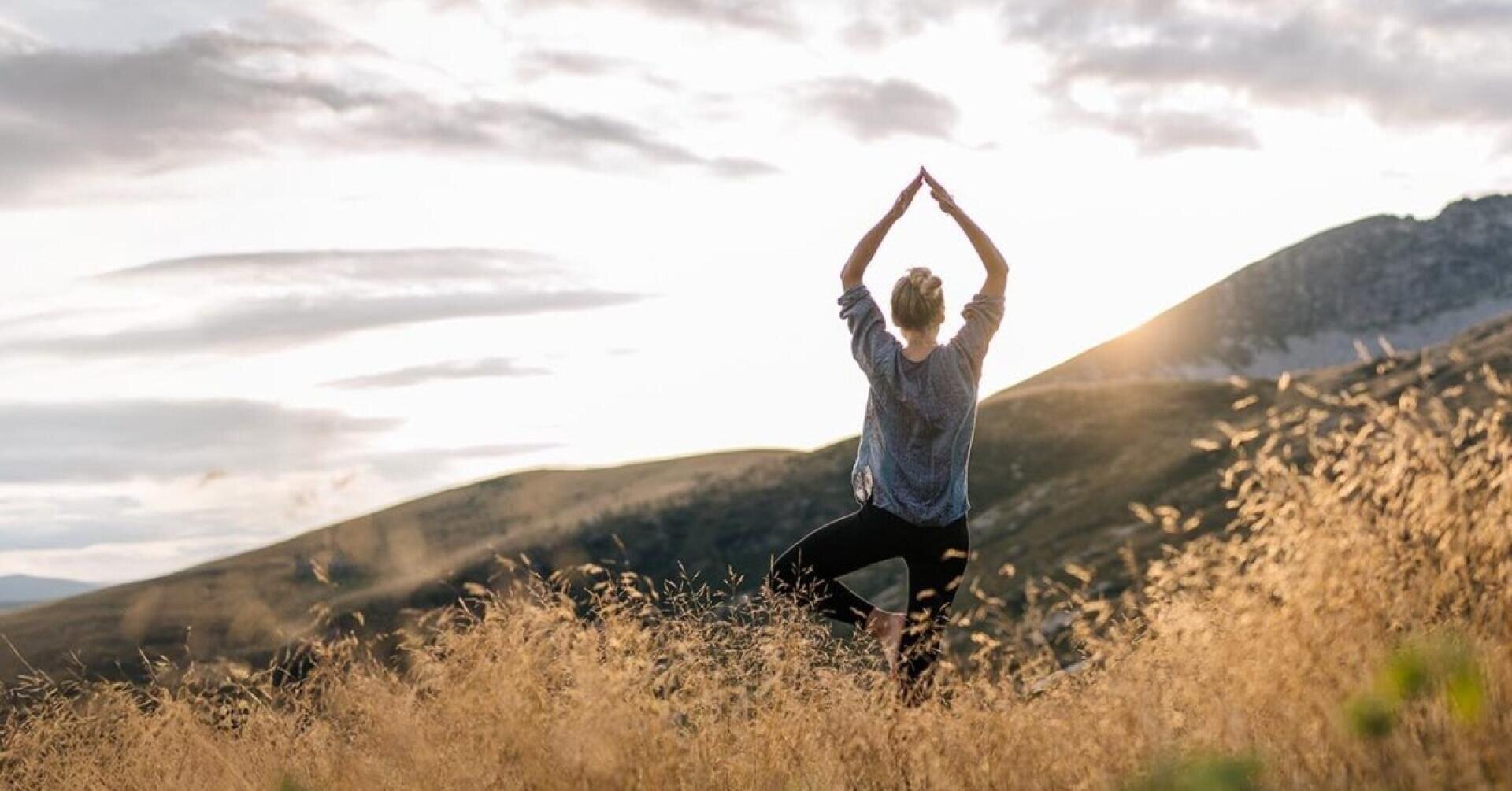 Eine Person steht in einer Yoga-Baum-Pose auf einem grasbewachsenen Hügel bei Sonnenuntergang, mit Bergen im Hintergrund und dem Himmel voller sanfter Wolken.