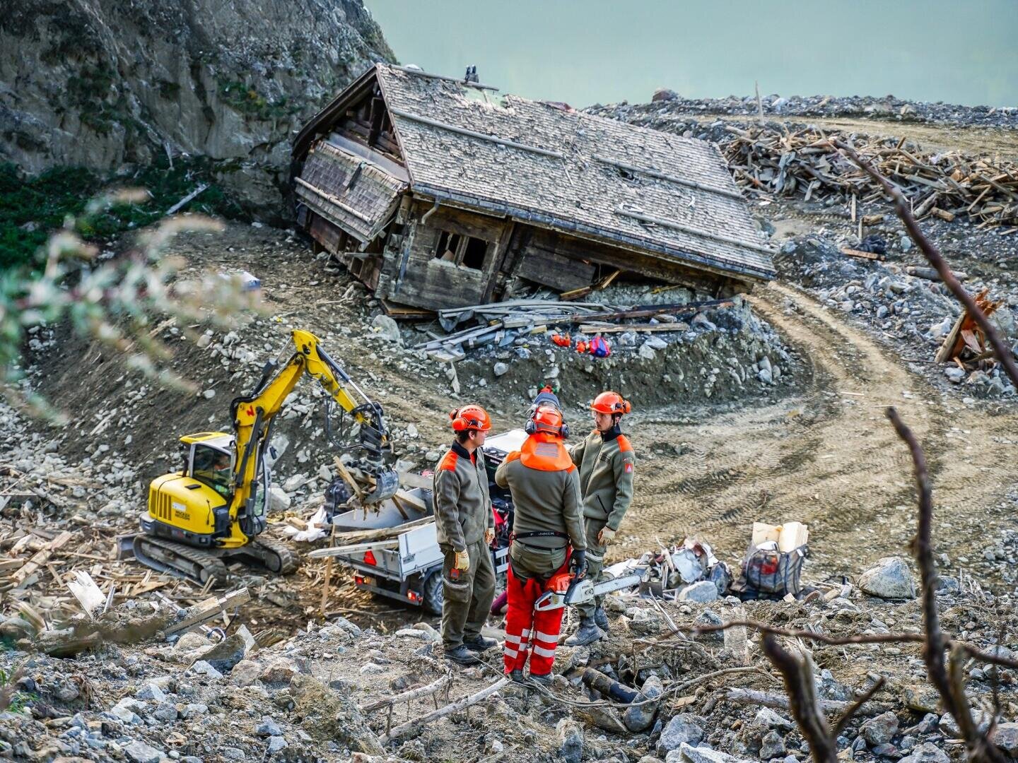 Rettungskräfte in Schutzausrüstung stehen in der Nähe von Geräten und einem kleinen Bagger an der Stelle eines Erdrutsches, an der ein beschädigtes Haus inmitten von Trümmern und felsigem Gelände auf die Seite gekippt ist.