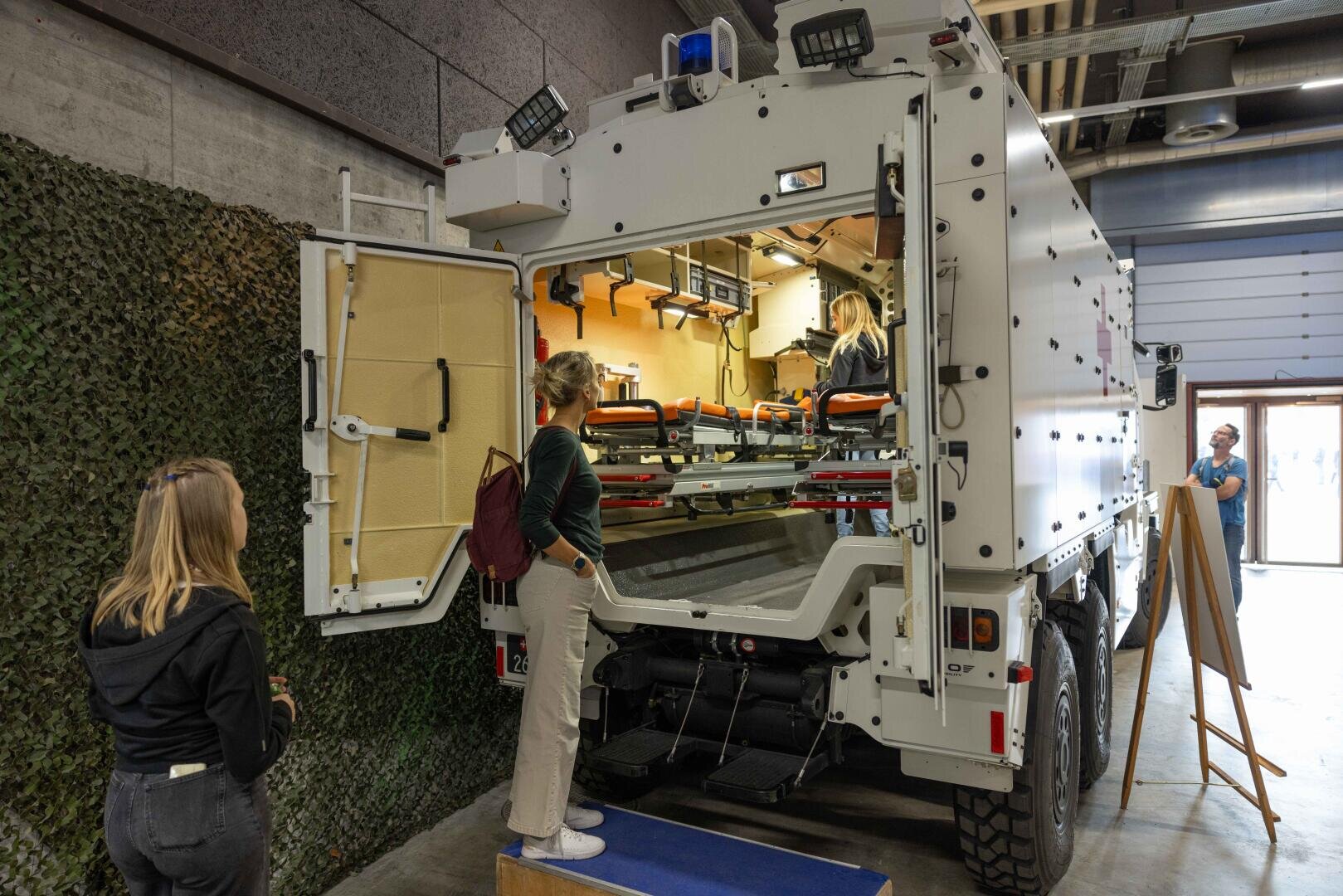 Zwei Frauen blicken in das offene Heck eines weißen, gepanzerten Sanitätsfahrzeugs, das mit Tragen und medizinischer Ausrüstung ausgestattet ist, während eine Person darin sitzt. Die Szene findet in einem Innenraum statt, möglicherweise auf einer Ausstellung oder Veranstaltung.