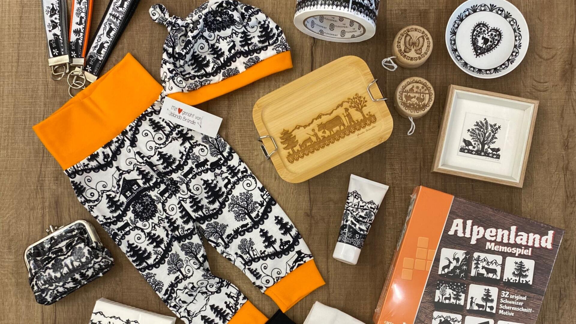Eine flache Anordnung von alpenländischen Gegenständen, darunter eine schwarz-weiß gemusterte Babyhose mit orangefarbenem Bündchen und Hut, eine Holzkiste, eine Kosmetiktube, ein Memory-Spiel, Geldbörsen, Schmuck und andere kleine Geschenke auf einer Holzfläche.