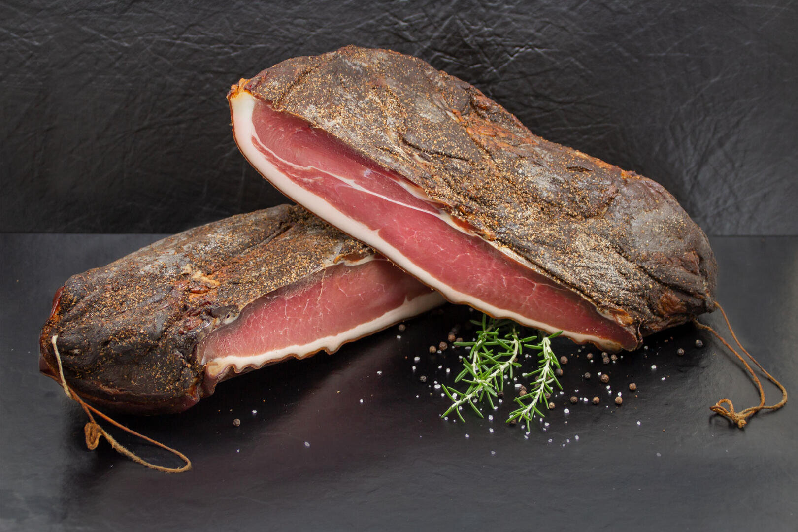 Zwei Stücke gepökeltes Fleisch mit dunklen, gewürzten Außenschichten sind auf einer schwarzen Fläche gestapelt. Vor dem Fleisch liegen ein kleiner Rosmarinzweig sowie einige Pfefferkörner und Salz gestreut.