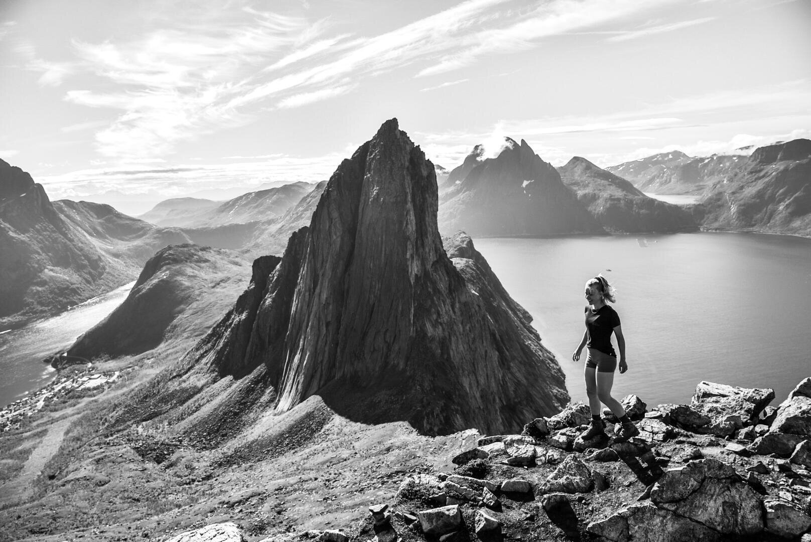 Eine Person steht auf einem felsigen Gebirgskamm mit Blick auf dramatische, zerklüftete Gipfel und einen See oder Fjord darunter, mit fernen Bergen unter einem teilweise bewölkten Himmel, in Schwarz und Weiß.