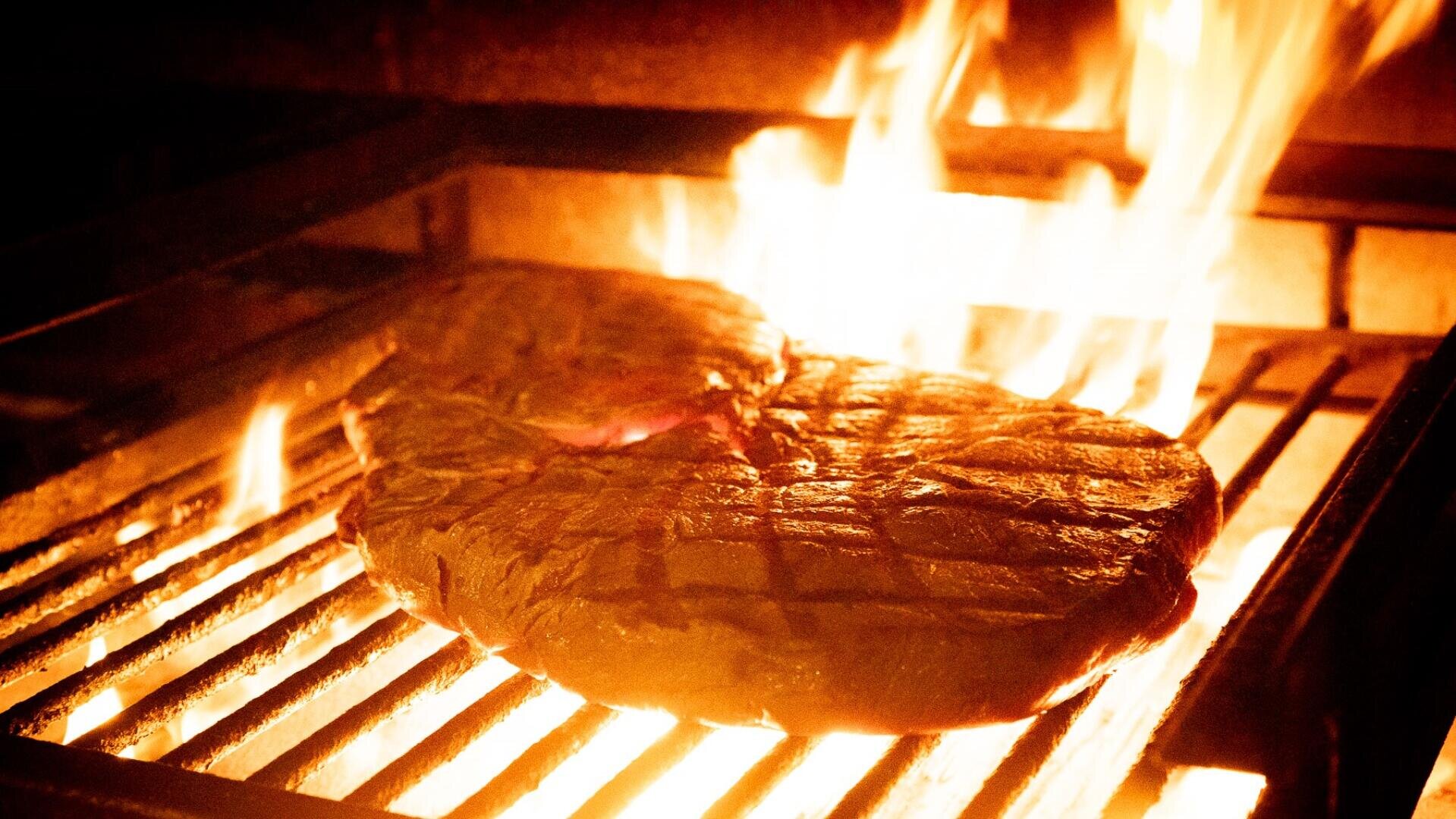 Ein dickes Steak wird über heißen Flammen auf einem Grill gegrillt, wobei das Feuer das Fleisch und den Grillrost beleuchtet.